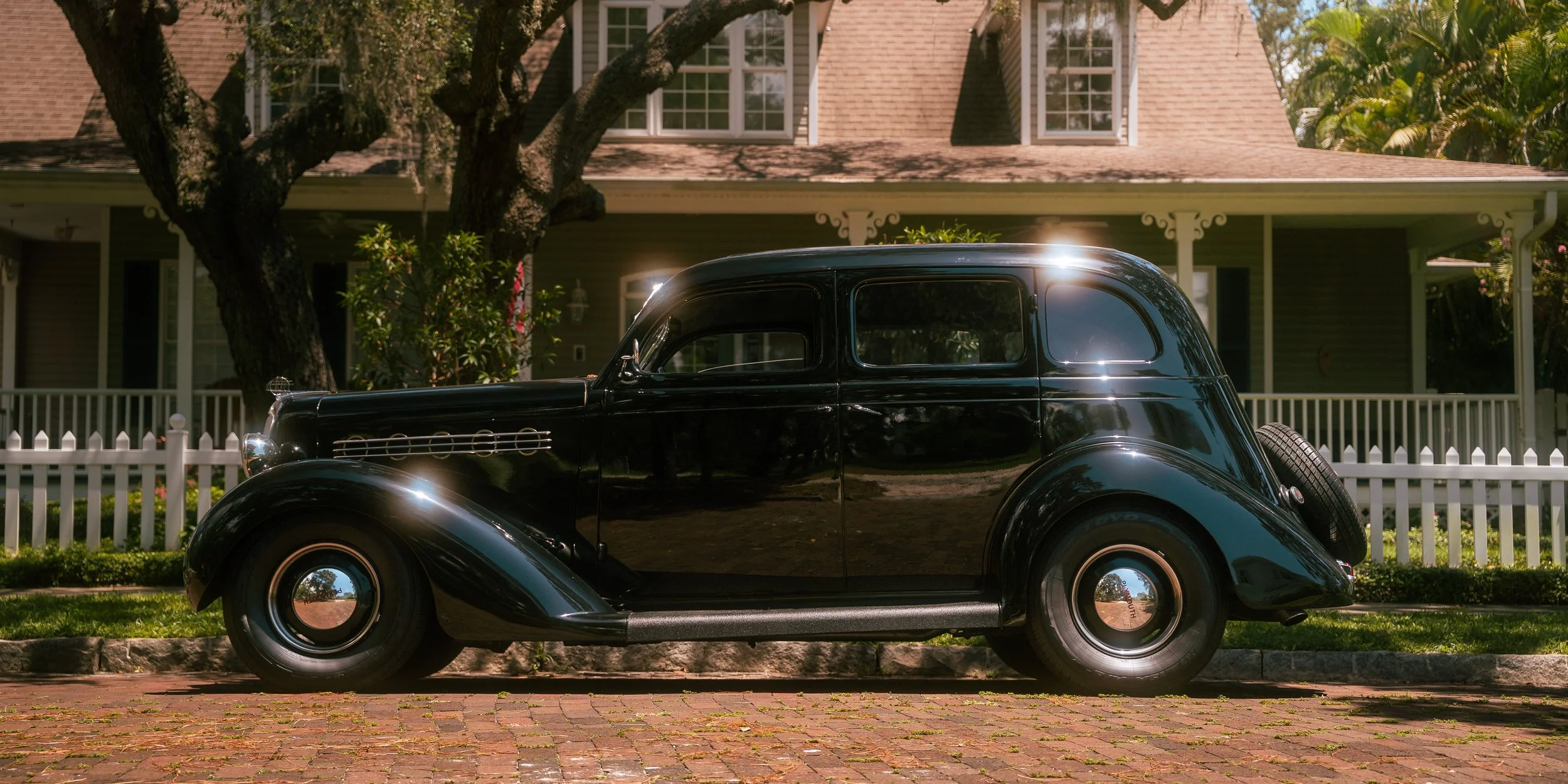 A vintage black car parked on a brick sidewalk in front of a house with a white picket fence and lush green trees.