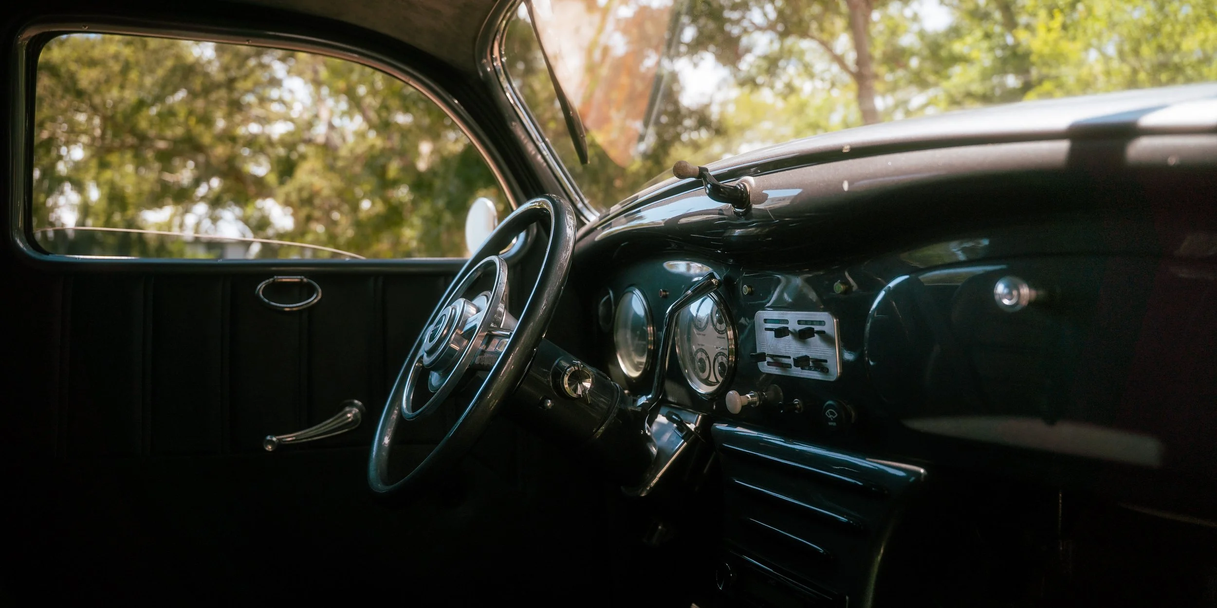 Interior of a vintage car showing the steering wheel, dashboard with round gauges, and minimalistic controls, with trees visible outside the window.