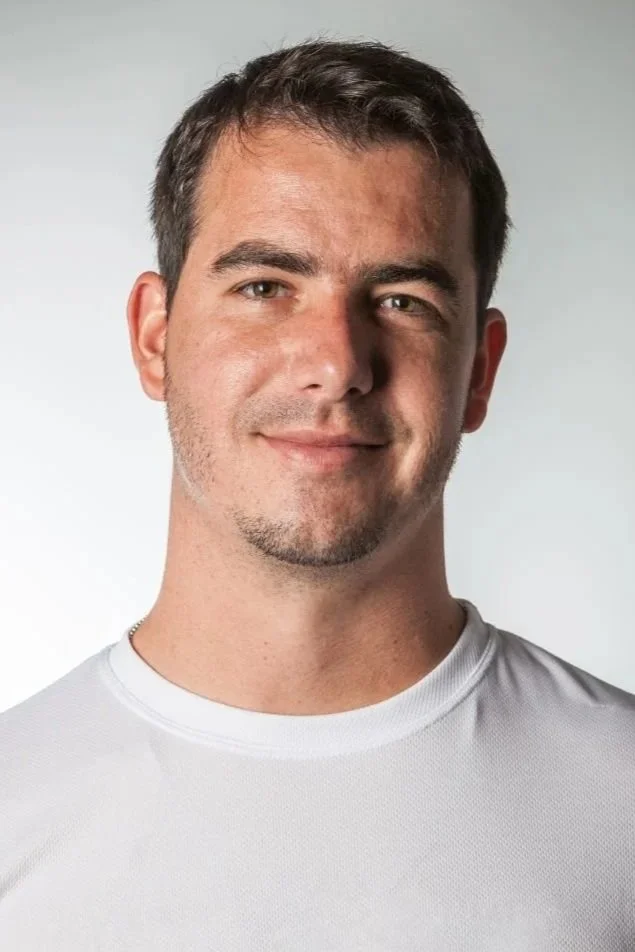 A young man with short brown hair, slight facial hair, and a light complexion, smiling, wearing a white shirt, against a neutral background.