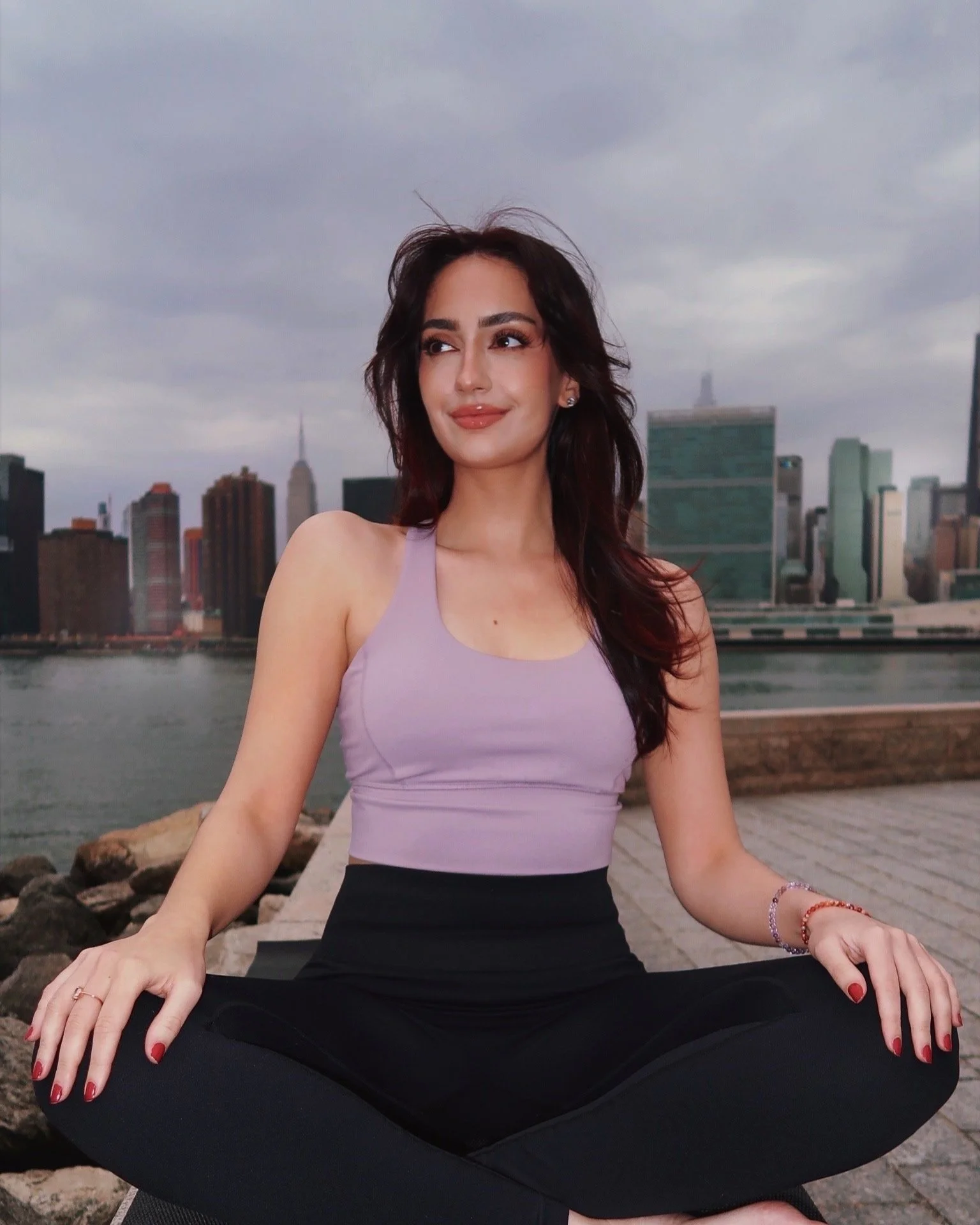 A woman practicing yoga by the river with a city skyline in the background.