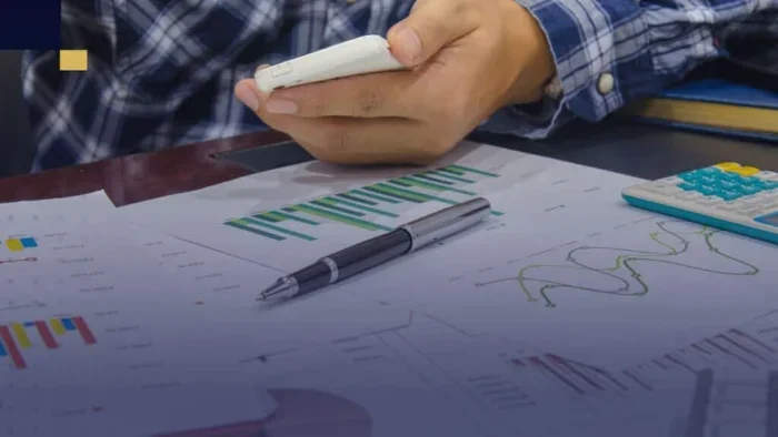 Mano sosteniendo un teléfono móvil sobre documentos y gráficas en una mesa.