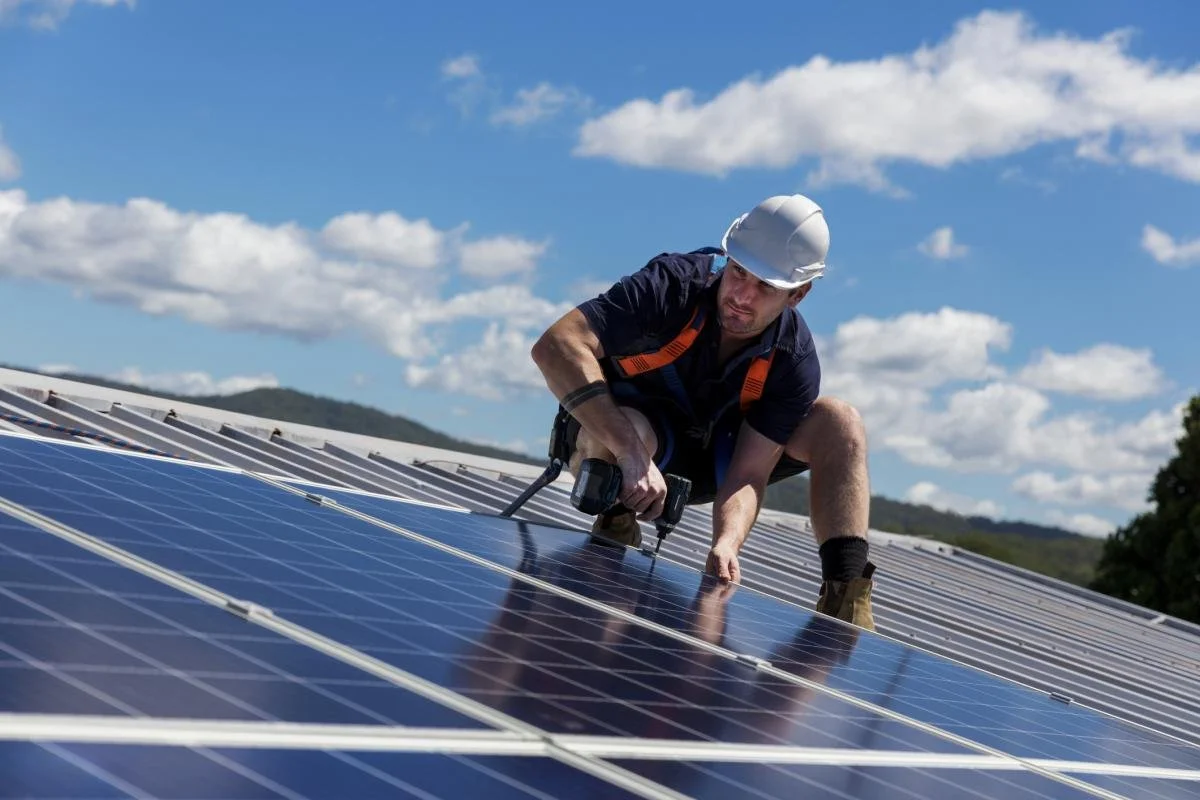 Un hombre con casco trabajando en paneles solares al aire libre bajo un cielo soleado con nubes.