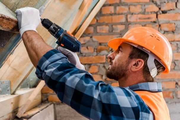 Hombre con casco naranja usando una taladro para trabajo en construcción de madera, en un muro de ladrillos.
