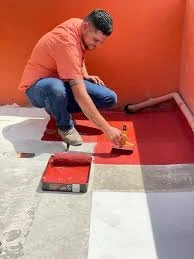Persona pintando un piso con pintura gris, en una habitación con paredes naranjas.