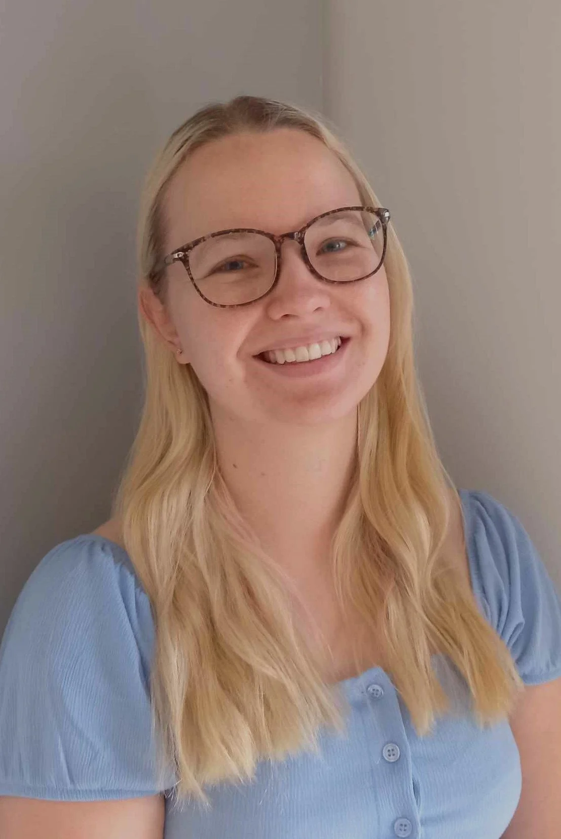 A young woman with long blonde hair, glasses, and a light blue button-up shirt smiling at the camera against a neutral-colored wall.