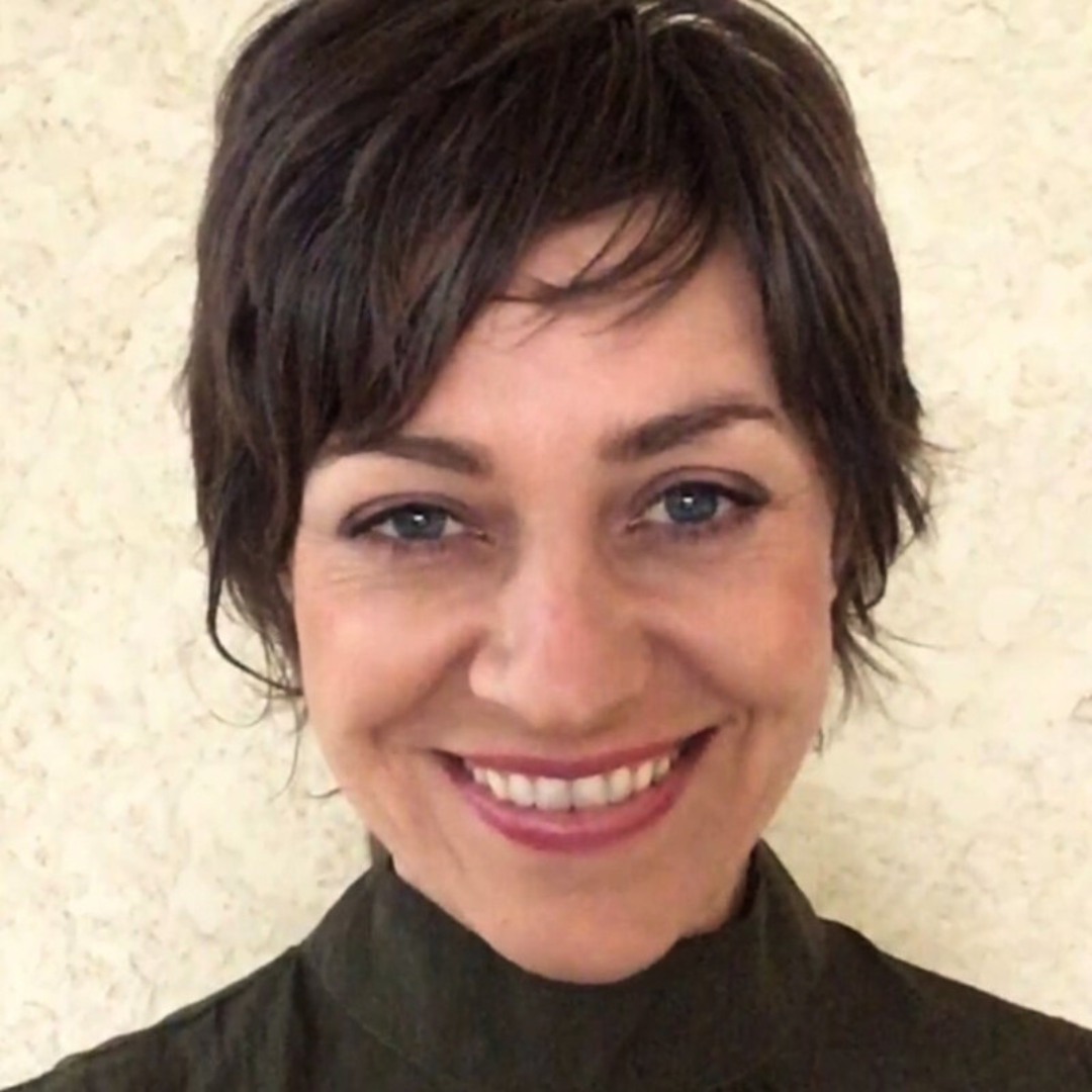 Smiling woman with short brown hair and blue eyes, wearing a black top, standing against a light-colored wall.