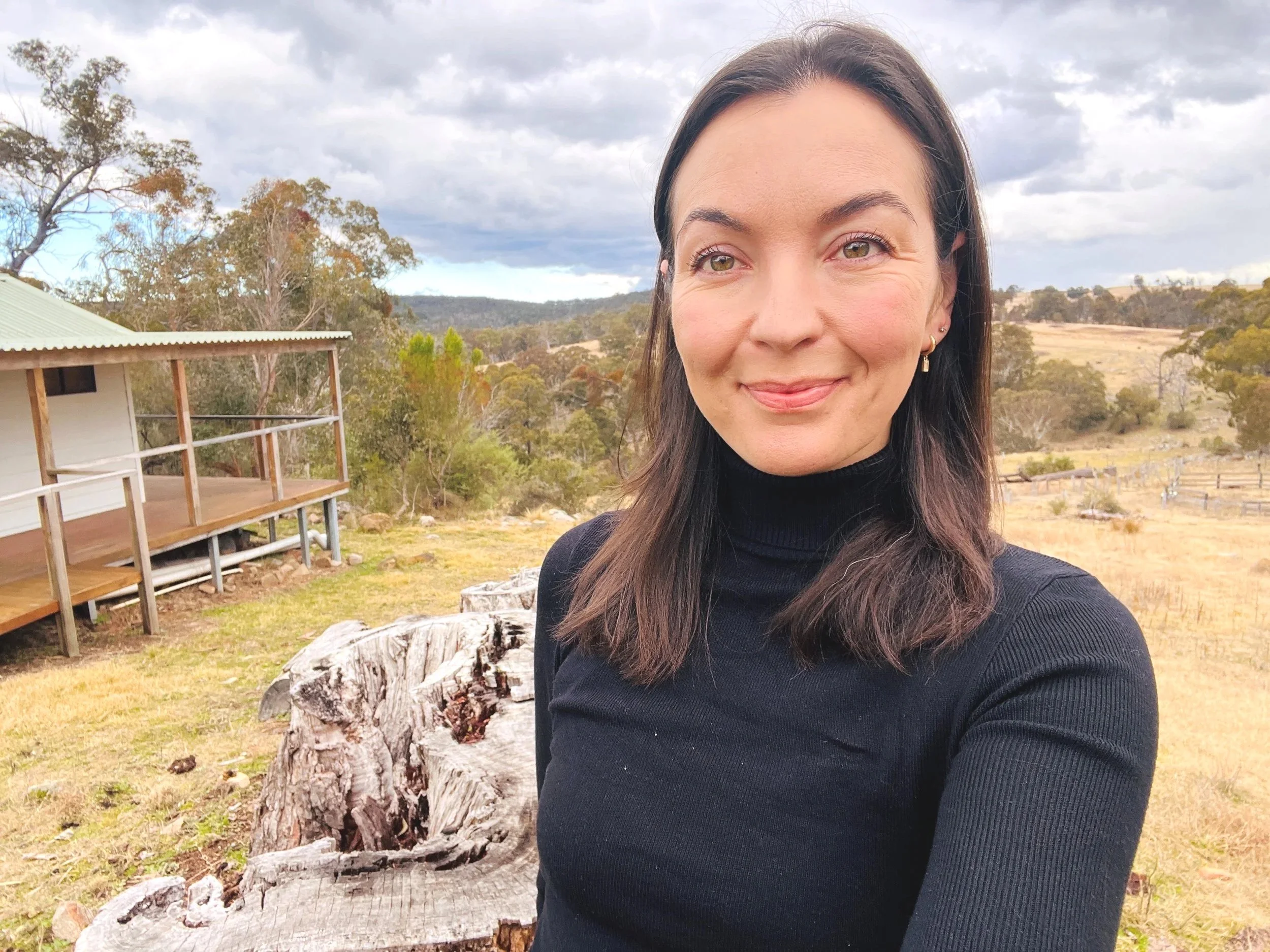 Close-up of a smiling woman with dark brown hair wearing a black turtleneck, outdoors in a rural setting with trees, rolling hills, and a cloudy sky in the background.
