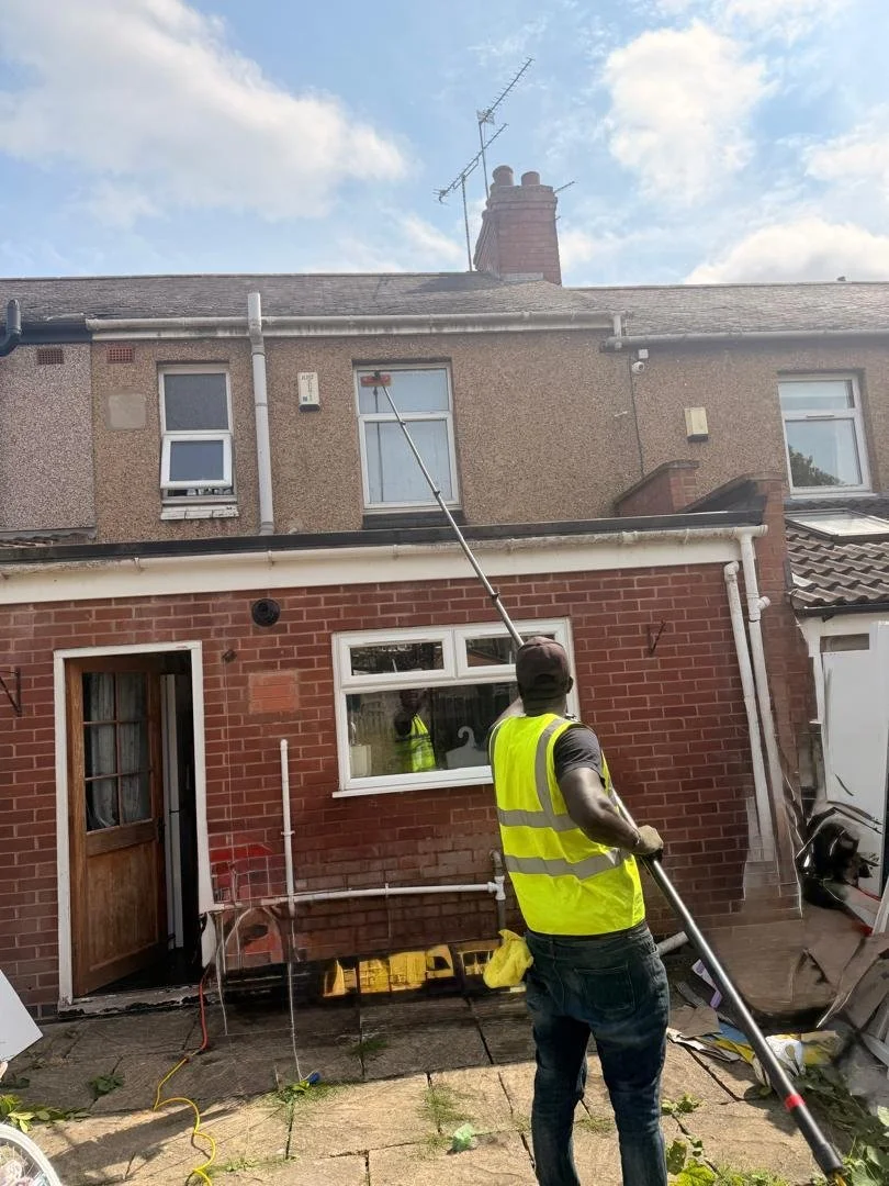 A worker in a yellow safety vest using a long pole to clean or reach the roof of a house with a brick and stucco exterior, in a residential neighborhood on a sunny day.