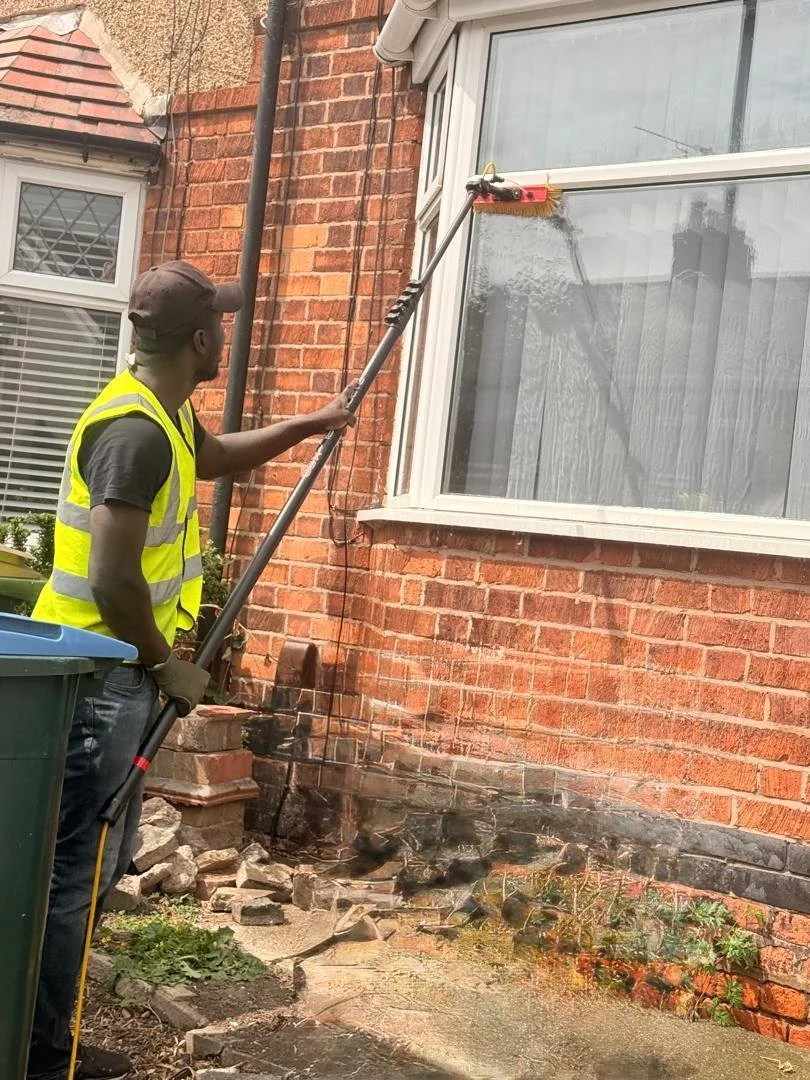 A man in a yellow safety vest using a pressure washer to clean the exterior brick wall of a house through a window.