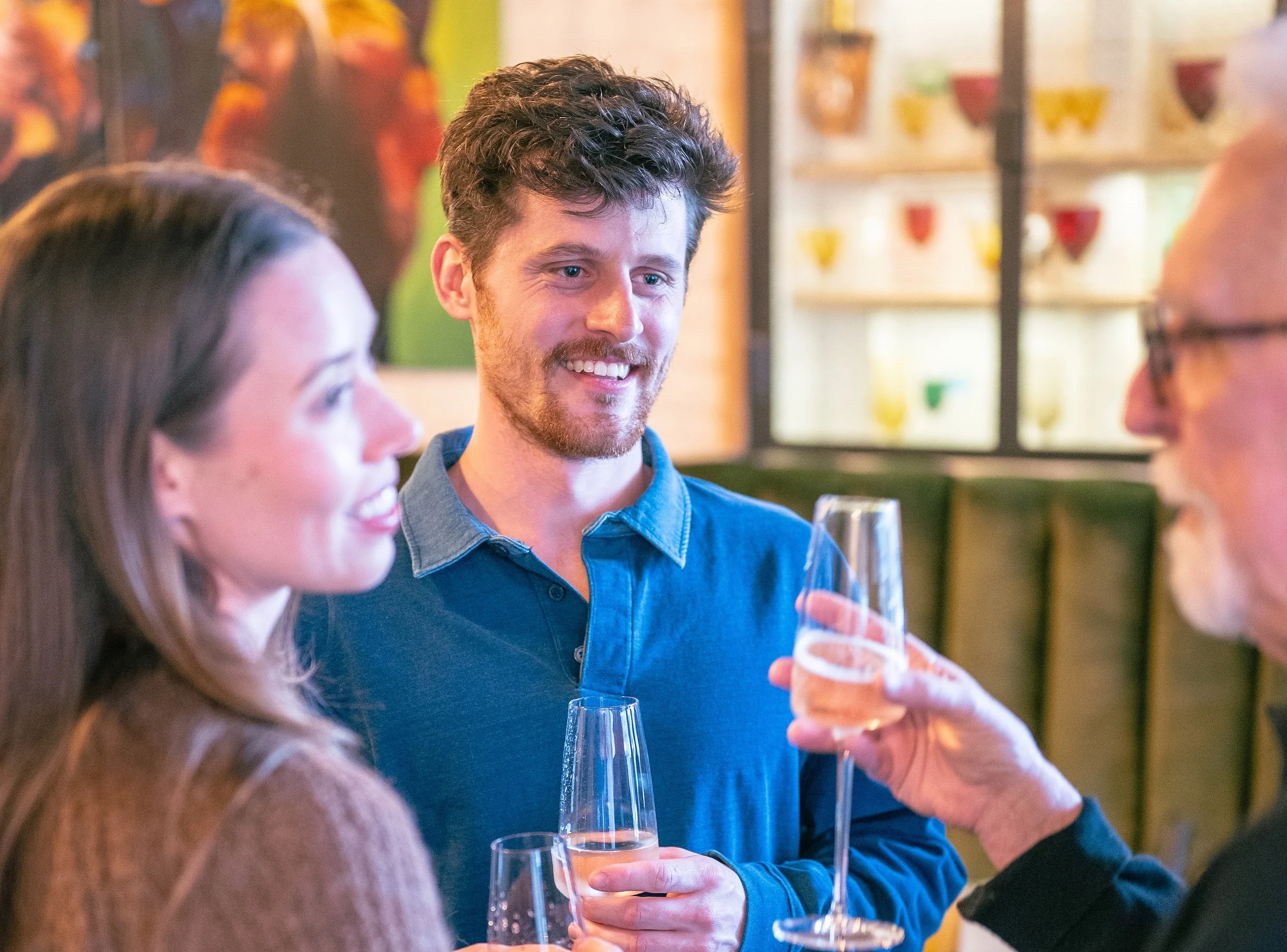 Three people having a conversation at a social gathering with drinks, in a warmly lit venue decorated with colorful art and shelves of glassware.