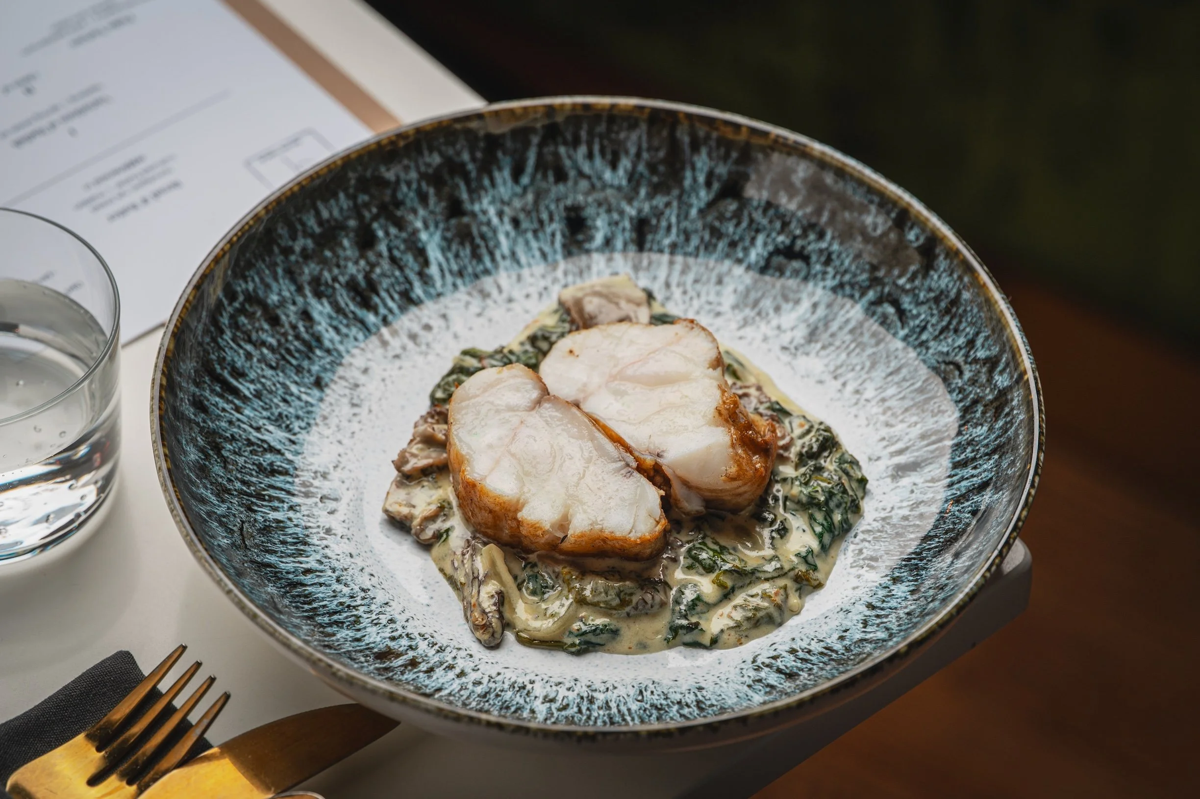 A bowl of cooked monkfish served over a creamy spinach sauce base, with a glass of water and a fork on a white table.