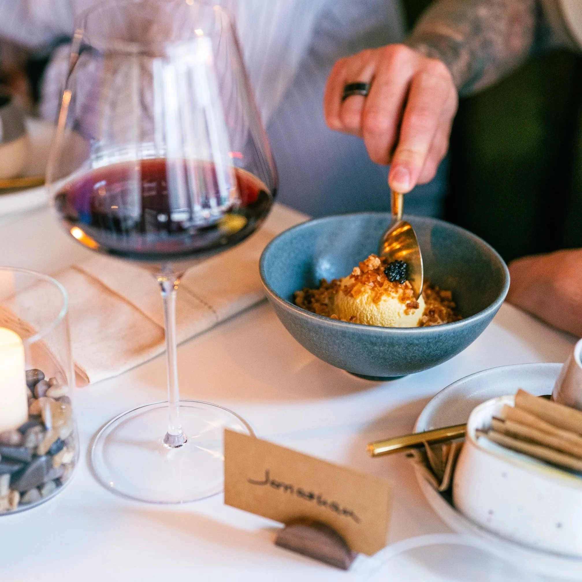 Close-up view of a glass of red wine, a person's hand holding a fork over a bowl of food, and other table items in the background.