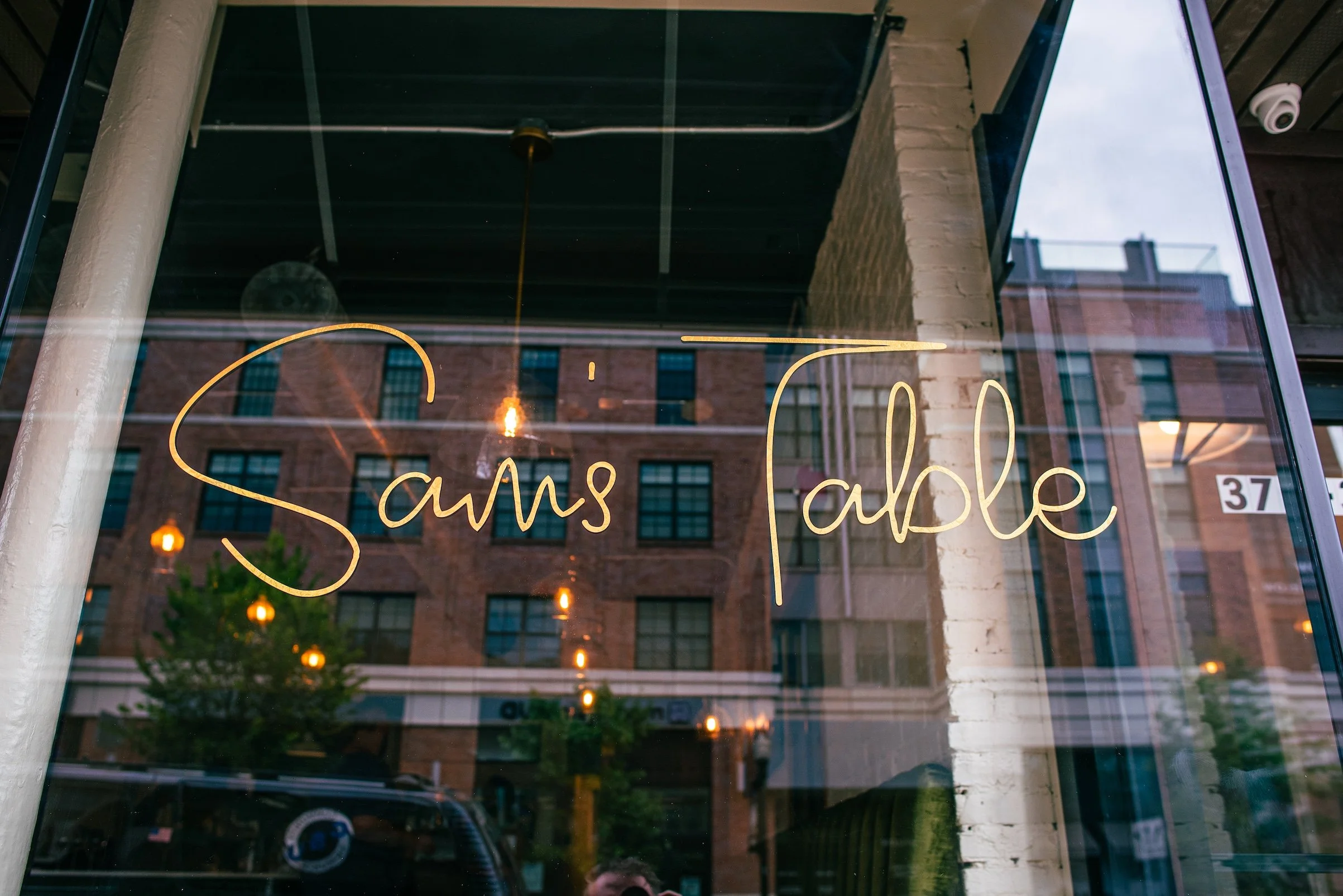 A glass storefront window with the words 'Sans Table' written in gold cursive lettering and reflections of brick buildings, trees, and hanging lights outside.