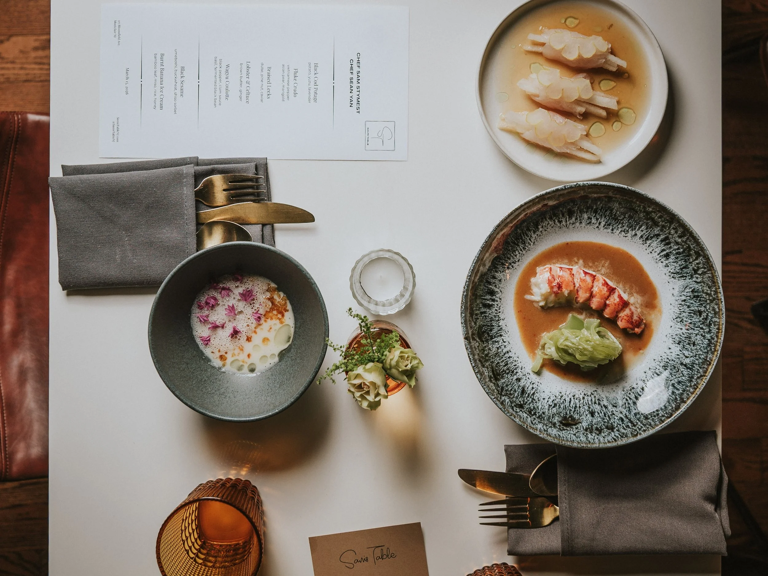 A white table set with a place setting that includes a gray napkin, gold fork and knife, several dishes of Asian cuisine, a small candle holder, and a printed menu. The dishes include a bowl of soup with pink flower petals, a bowl of or broth with se