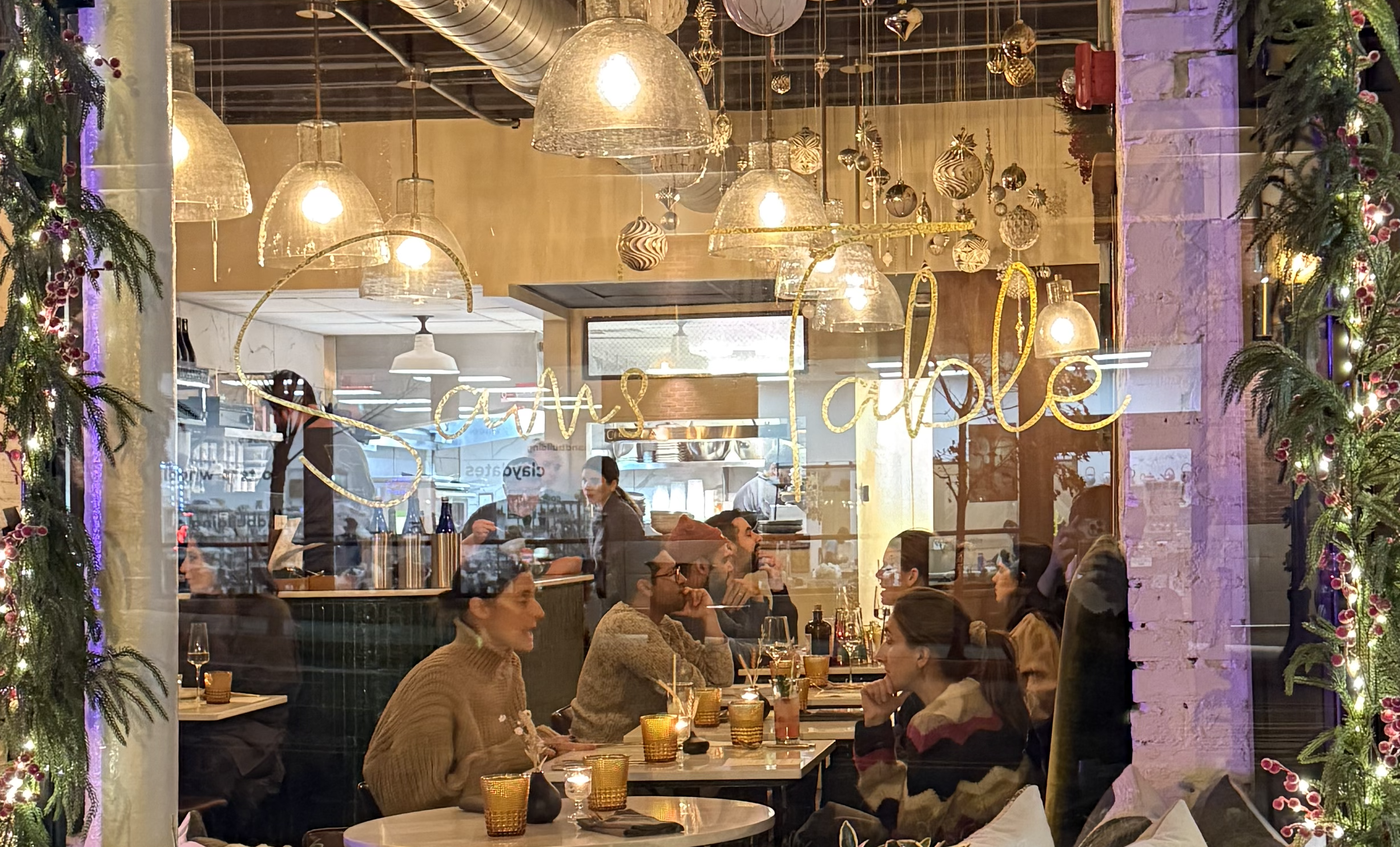 Sam's Table , a cozy restaurant decorated for Christmas, with hanging lights, festive ornaments, and guests dining at tables, seen through a glass window.