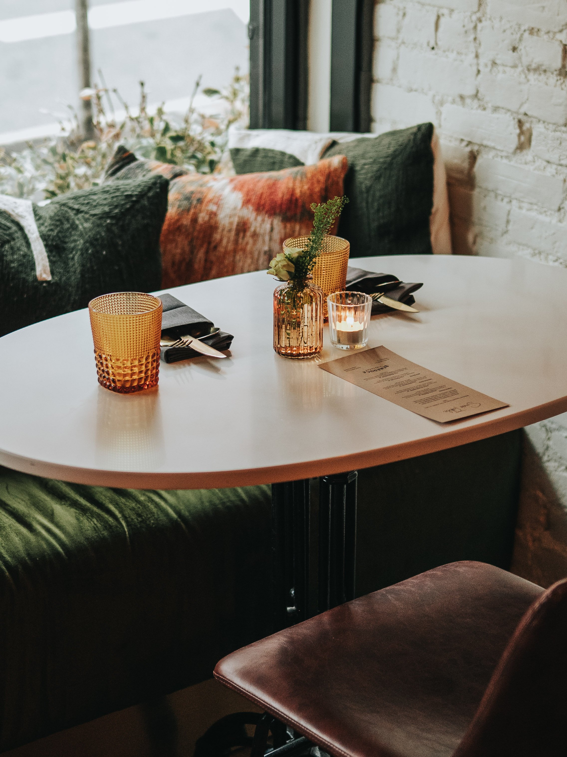Table setting in a cozy restaurant with a menu, two glasses, a candle in a decorative holder, and a small vase with greenery, against a backdrop of green cushioned seating and a white brick wall.
