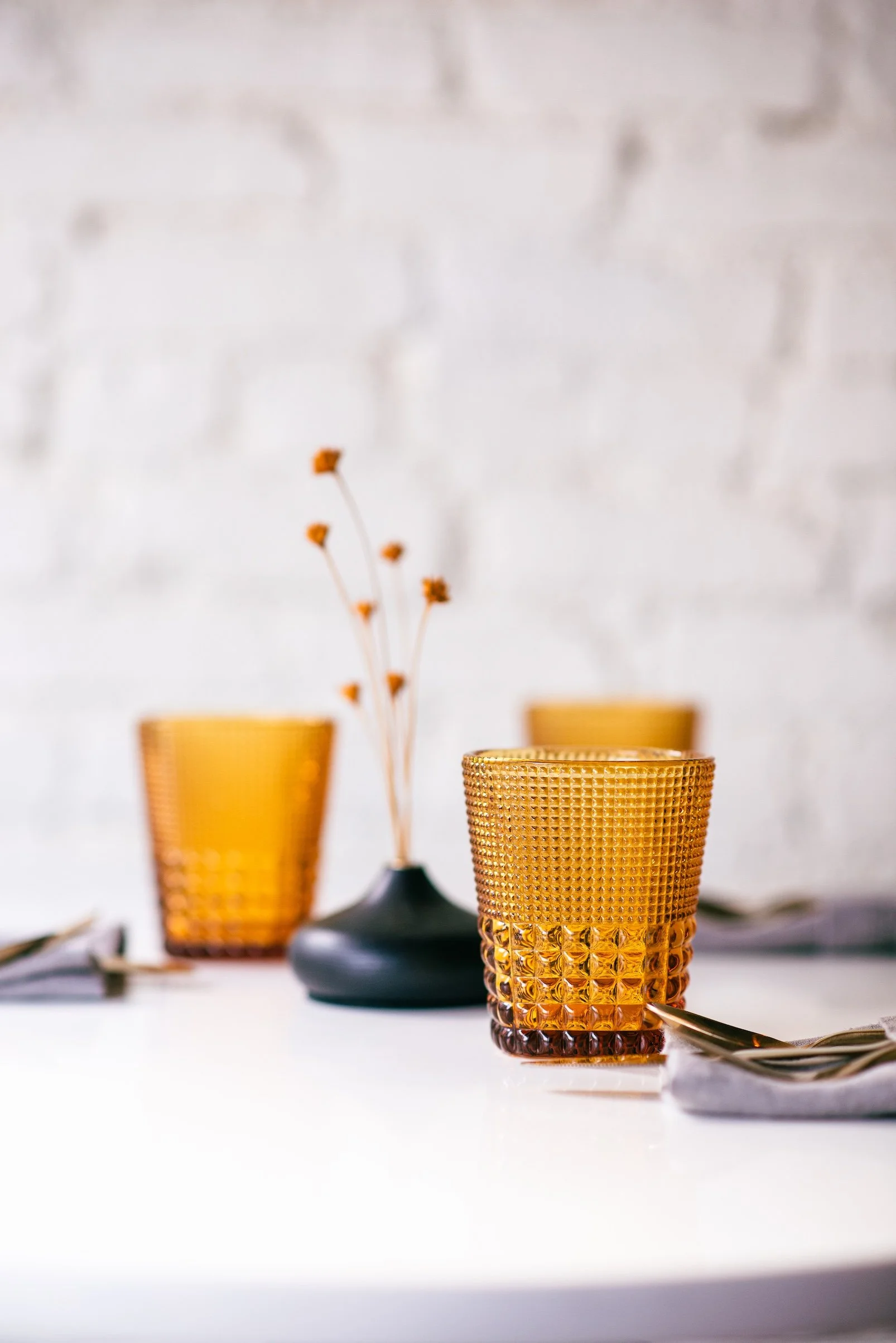 Inside Sam's Table, a set of three amber-textured glass cups on a white table, with a black vase holding dried flowers in the background, against a white brick wall.