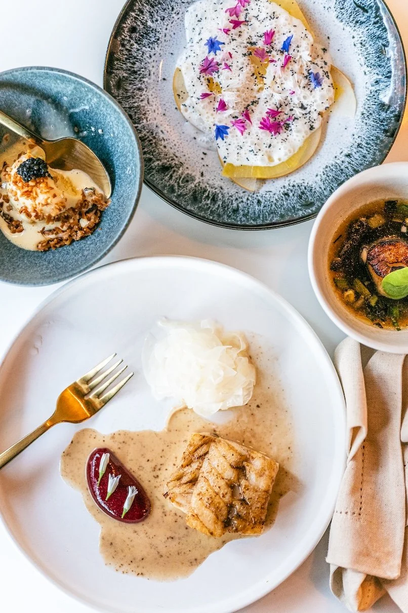 Assorted gourmet dishes including a plate with meat in sauce, whipped cream, and a garnished beet; smaller bowls with dessert, creamy cheese or yogurt with edible flowers, and a bowl of soup with green garnish.