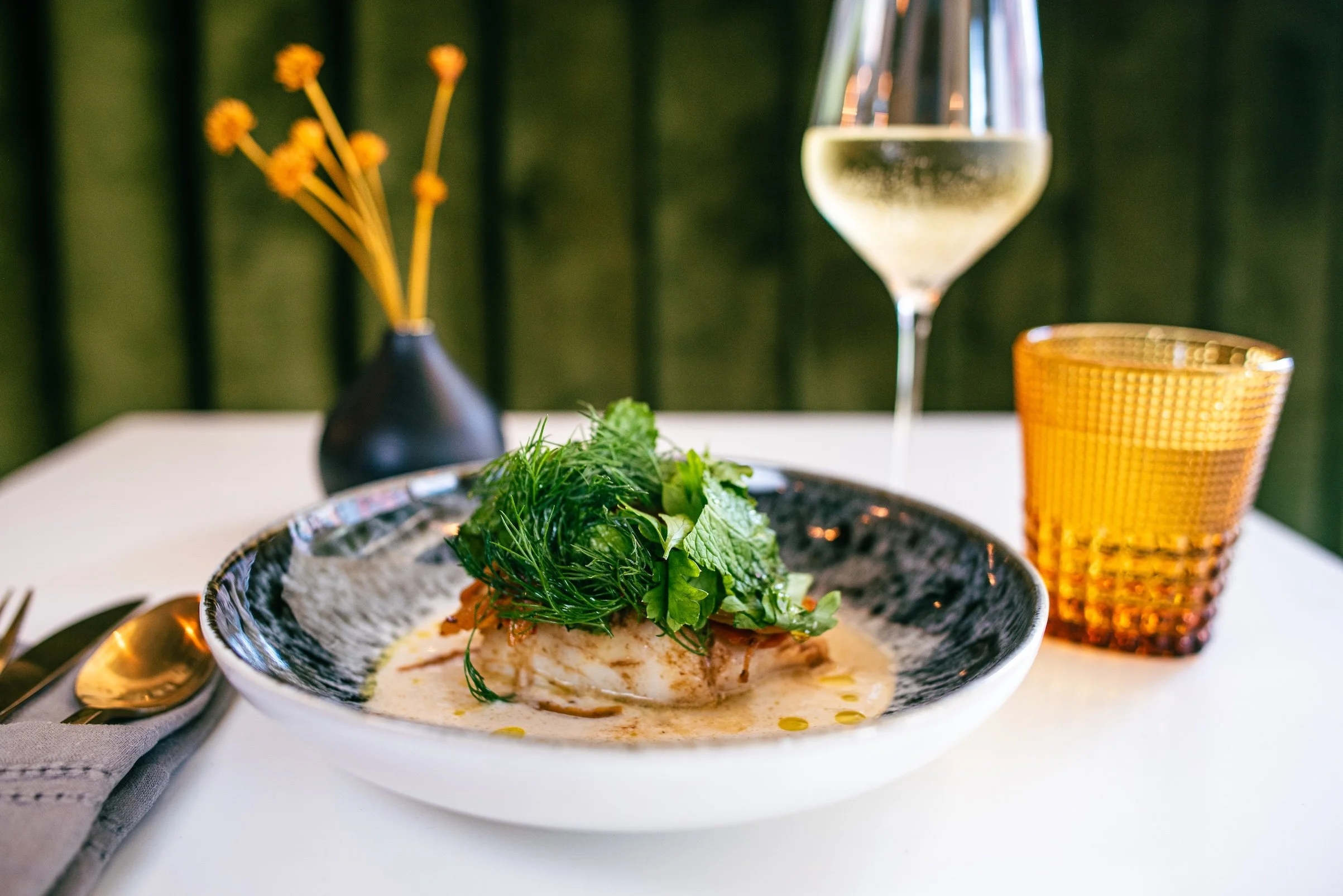 A plated fish dish with green herbs and sauce, a glass of white wine, and an orange glass of water on a white table with a black vase containing yellow flowers in the background.