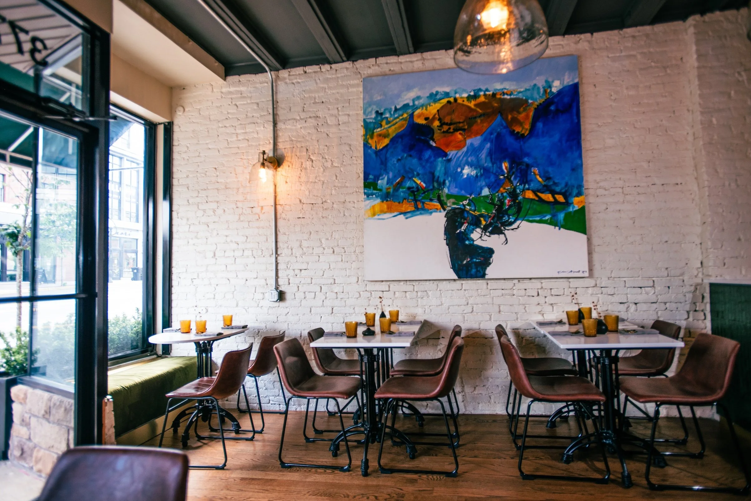 Sam's Table is a modern restaurant with white brick walls, colorful abstract artwork, brown leather chairs, white tables set with yellow glasses, and large windows letting in natural light.