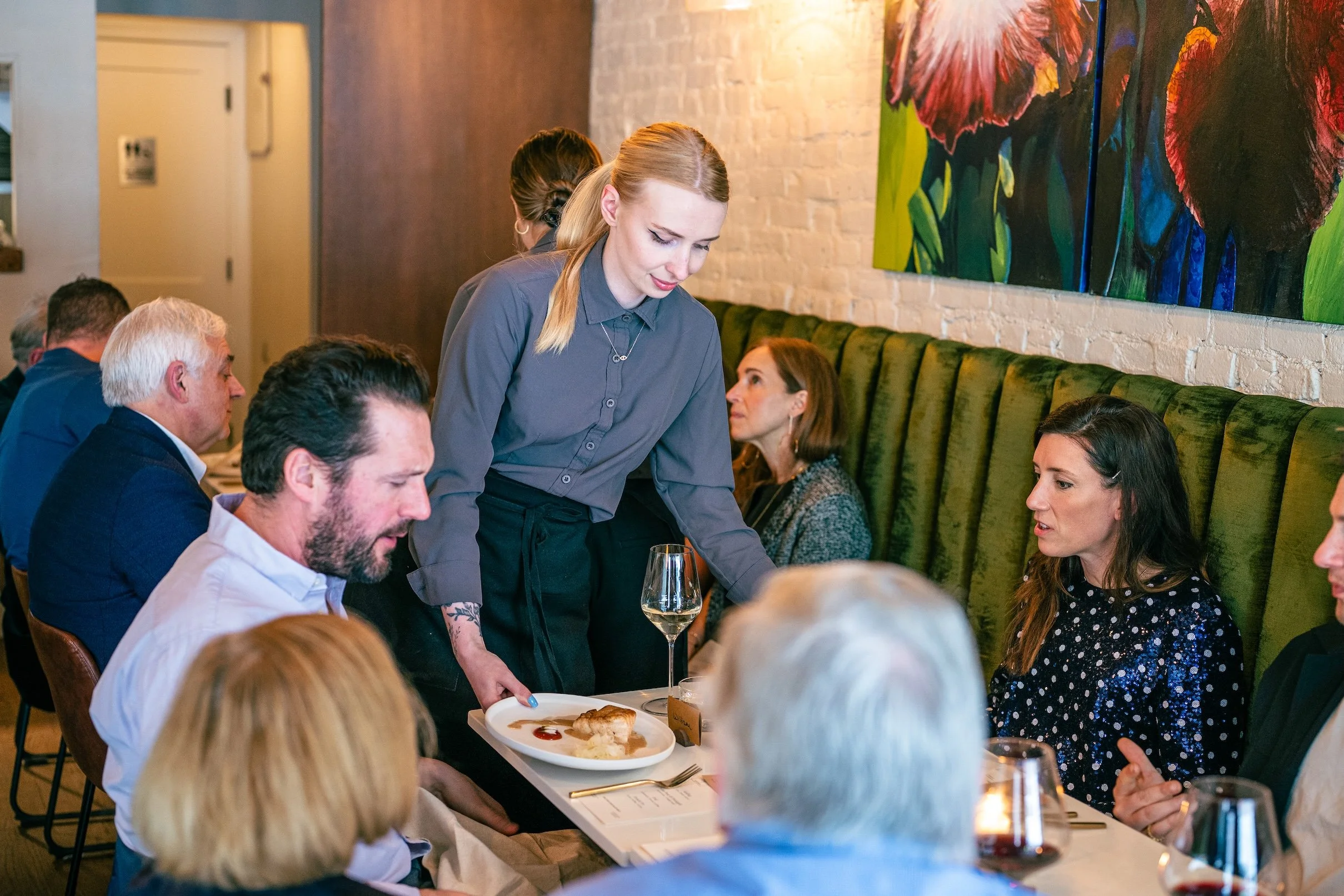 A woman with long blonde hair, wearing a grey shirt, serving food or drinks to a group of diverse people seated at a long dining table in a restaurant.