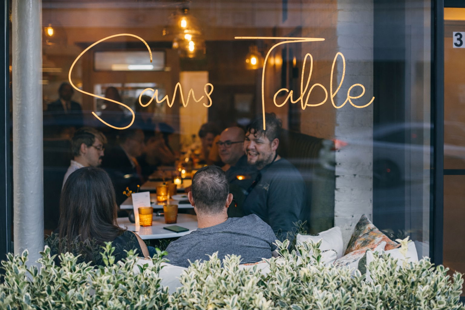 People dining inside a restaurant called 'Sams Table', seen through a glass window. The scene includes several guests talking and enjoying their meal, with warm lighting and a cozy atmosphere.