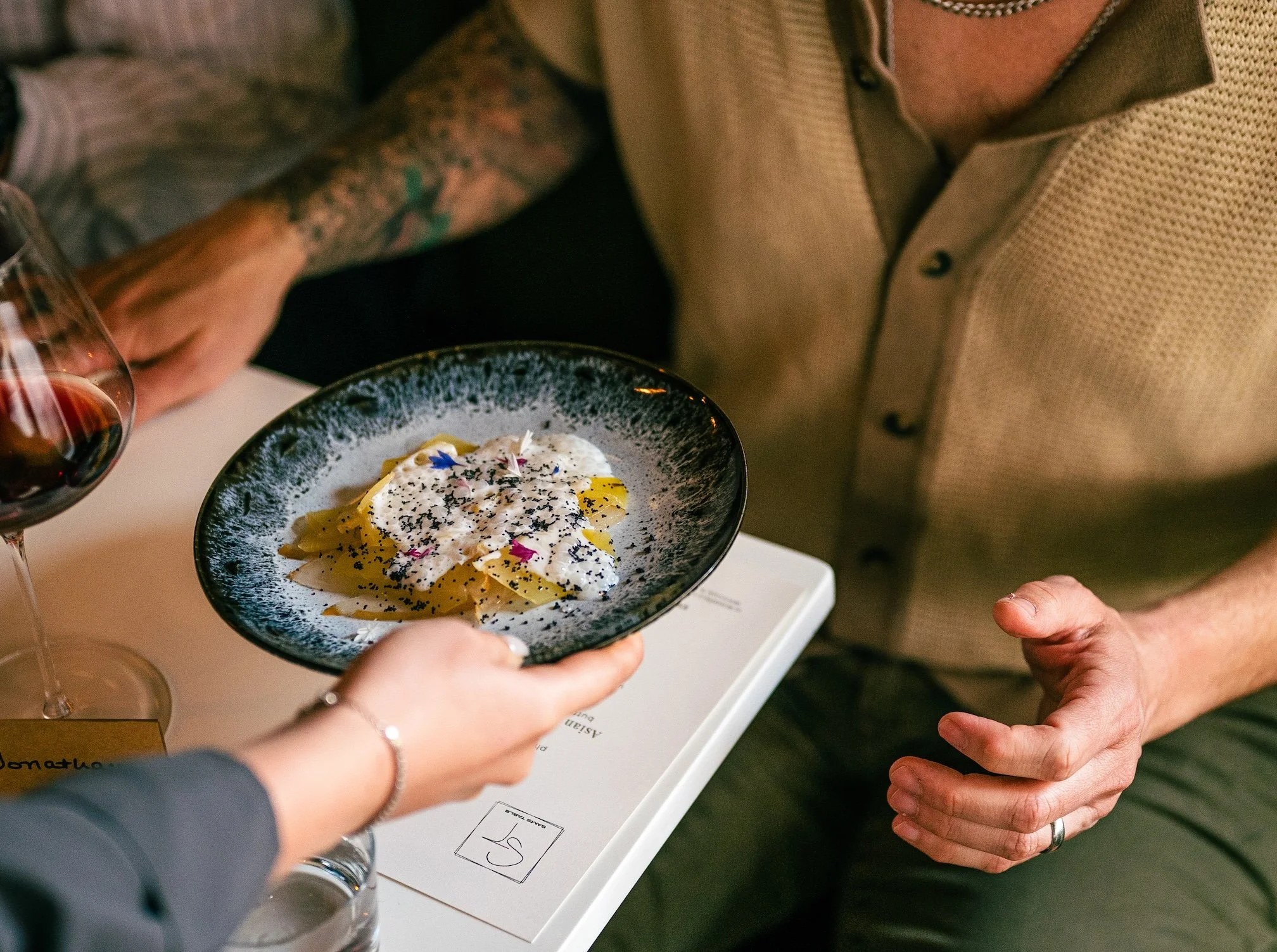 People exchanging a dish of pasta with cream sauce and garnishes at a restaurant, with a glass of red wine visible.