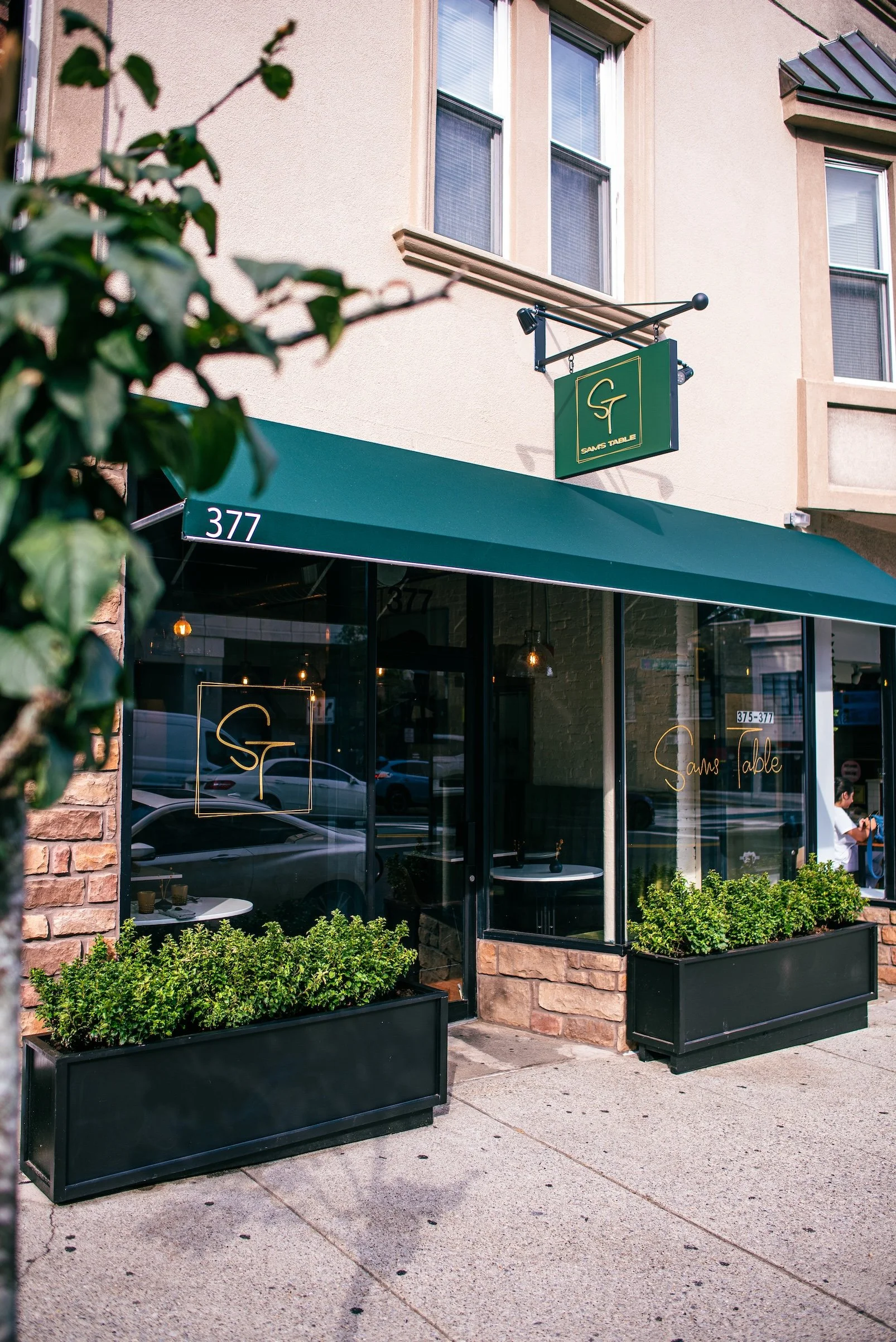 Sam's Table storefront features a green awning, large glass windows, and small black planter boxes with green bushes is located at 377 Bloomfield Avenue, Montclair NJ