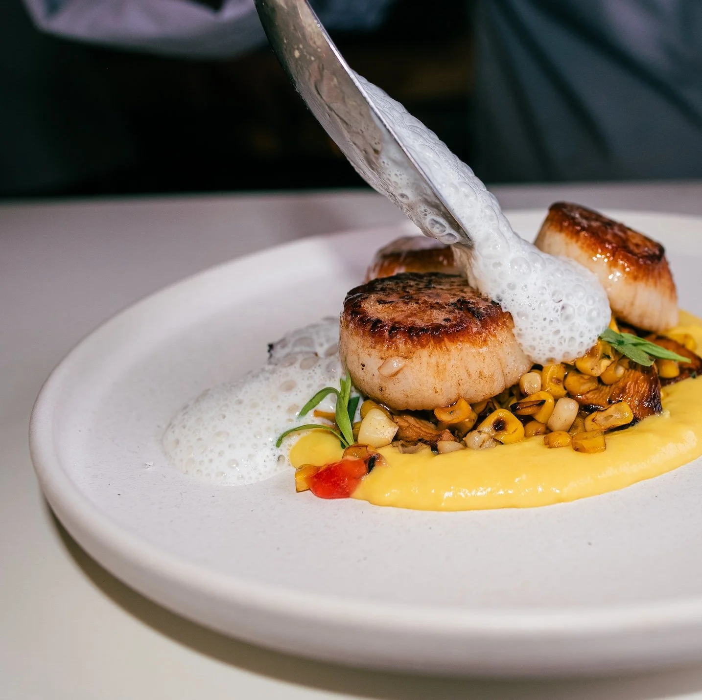Seared scallops served with corn, yellow sauce, and foam on a white plate.