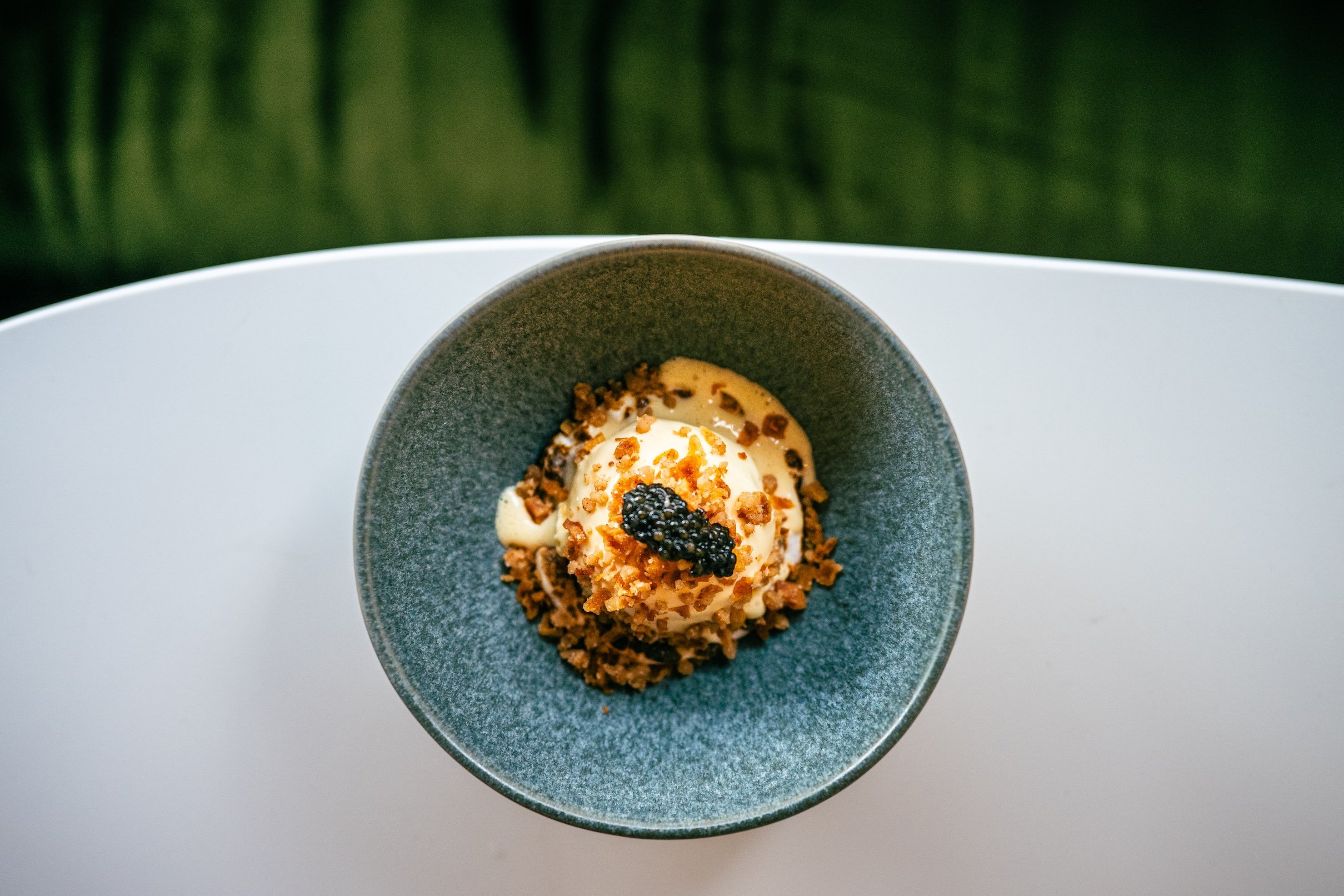 Dessert with ice cream, black caviar, and crunchy toppings served in a gray dish, placed on a white surface with a blurred green background.