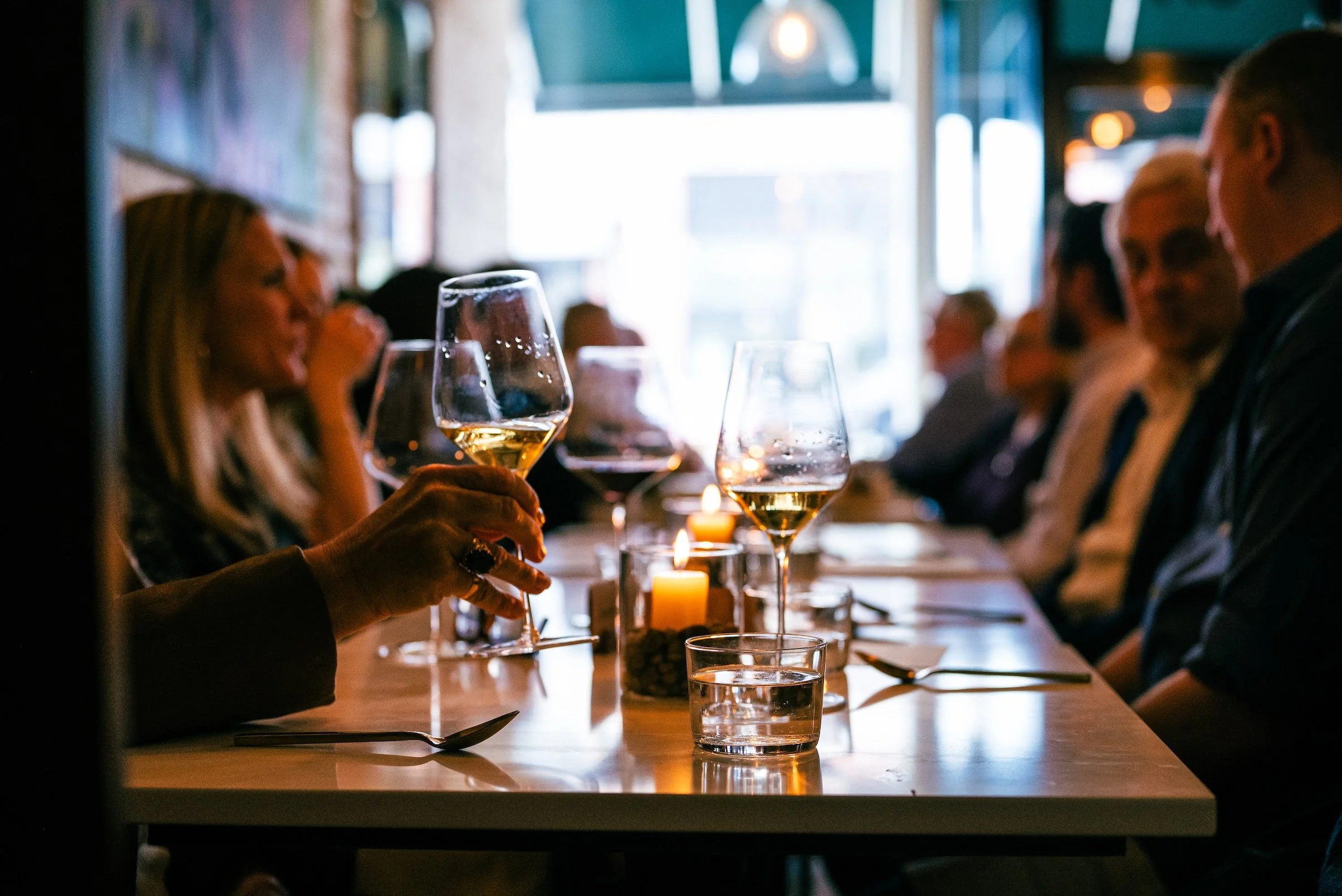 People sitting at a table in a dimly lit restaurant, with glasses of white wine, candles, and a bright window in the background.