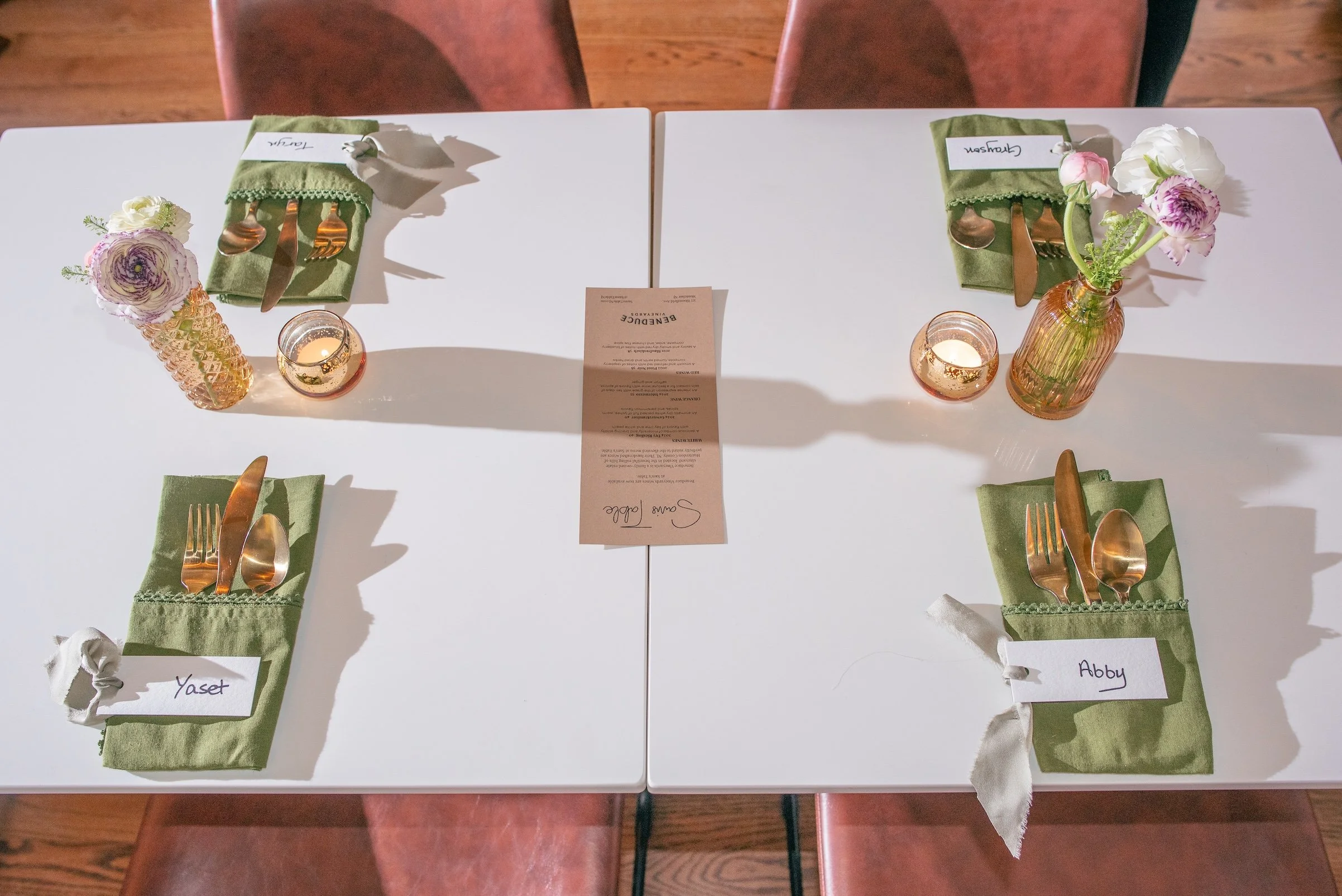 Two tables set for a meal with green napkins, gold-colored cutlery, small votive candles, and small flower arrangements in amber and orange vases, labeled with guest names Yasmin, Abby, and Clayson. A menu card is centered between the tables.