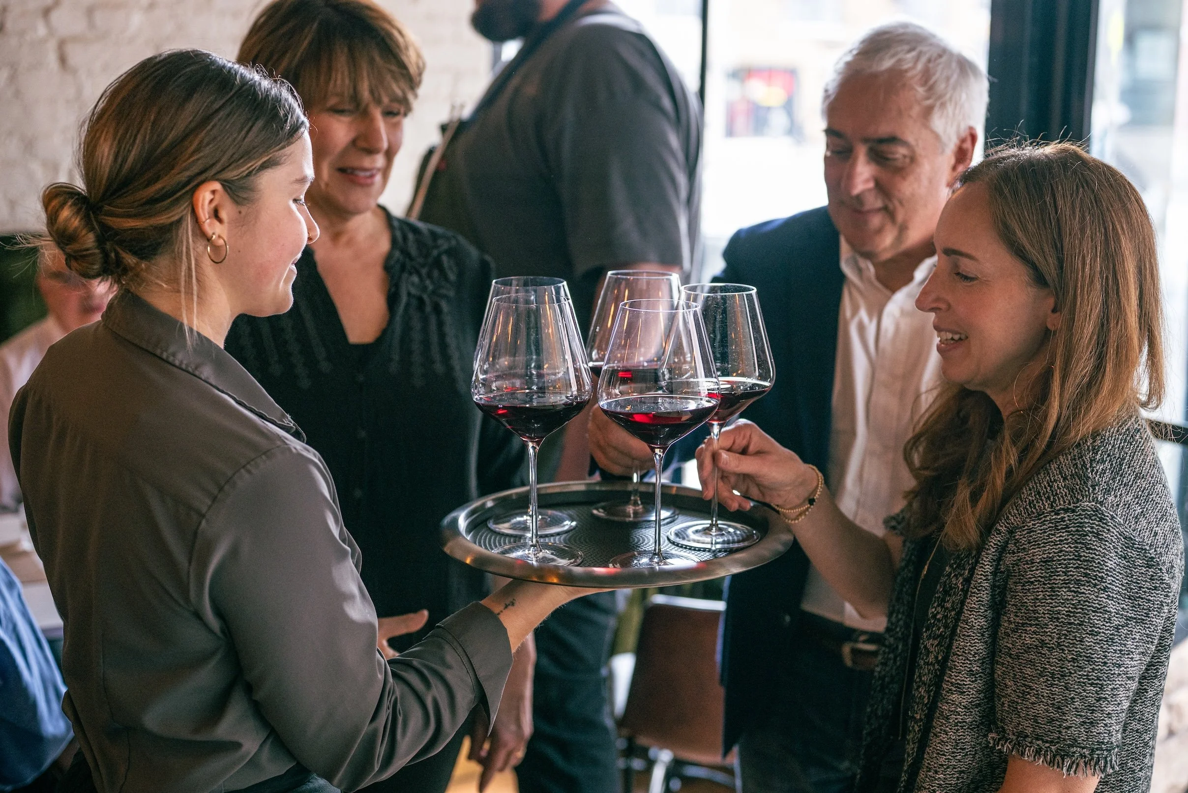 A waitress serving three glasses of red wine to a woman in a restaurant with other customers and waitstaff visible in the background.