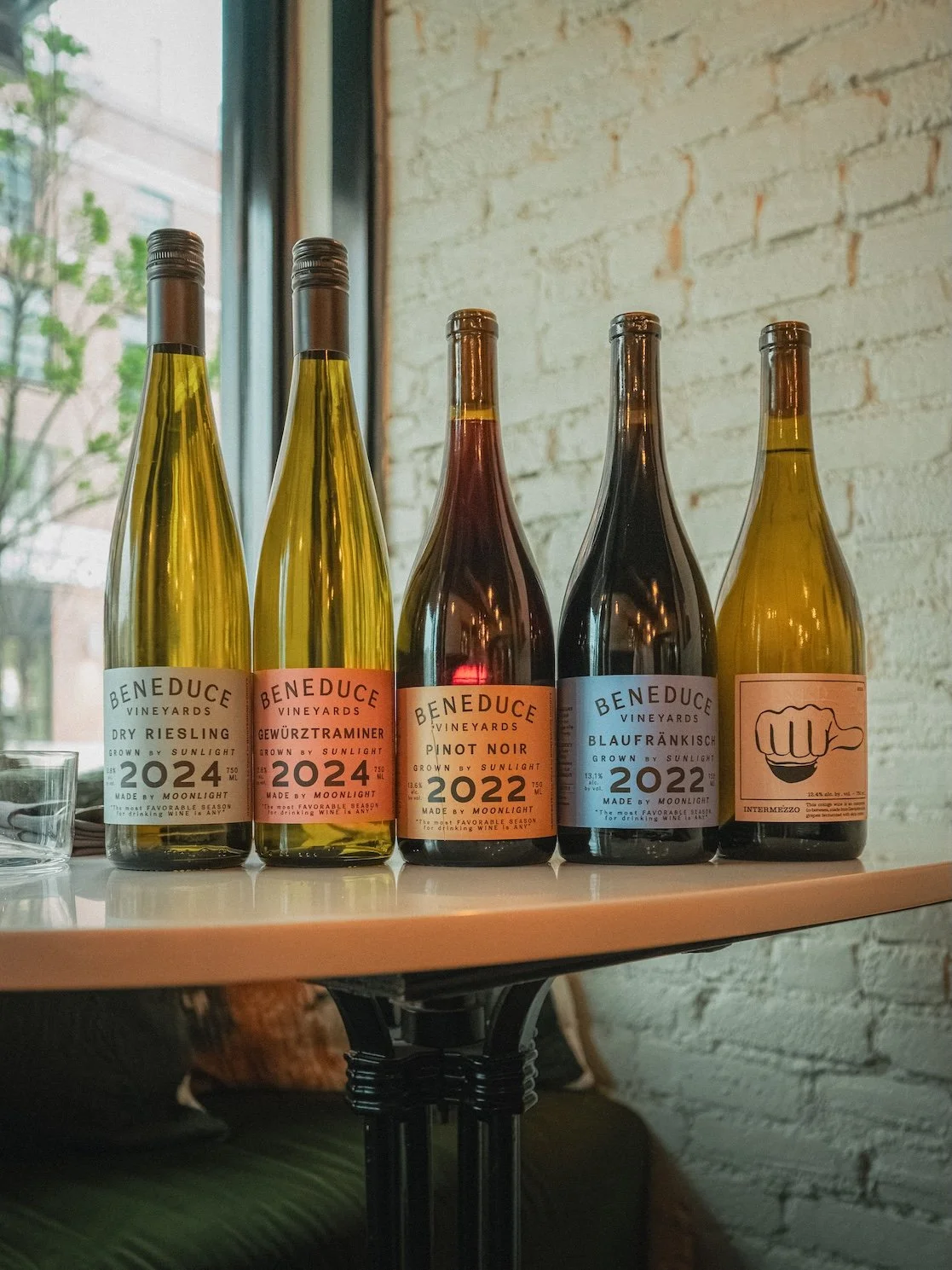 Beneduce Vineyards wines  on a table against a white brick wall, including Riesling, Gewurztraminer, Pinot Noir, Blaufrankisch, and an artful bottle with a fist logo.