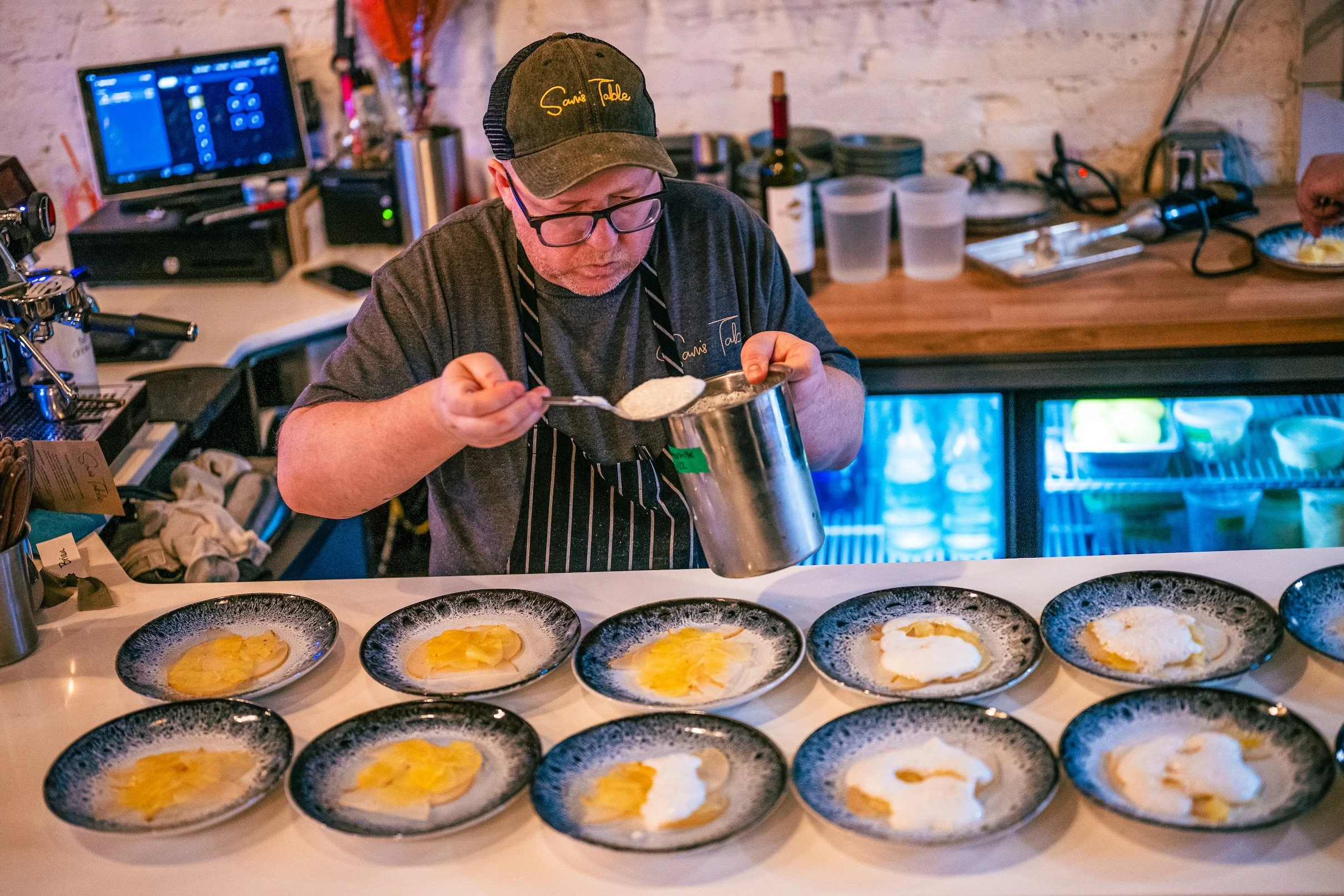 Chef Josh Pierson preparing plating an elegant meal at the counter. The chef is wearing glasses, a dark apron, and a cap with the embroidered text 'Sams Table'.