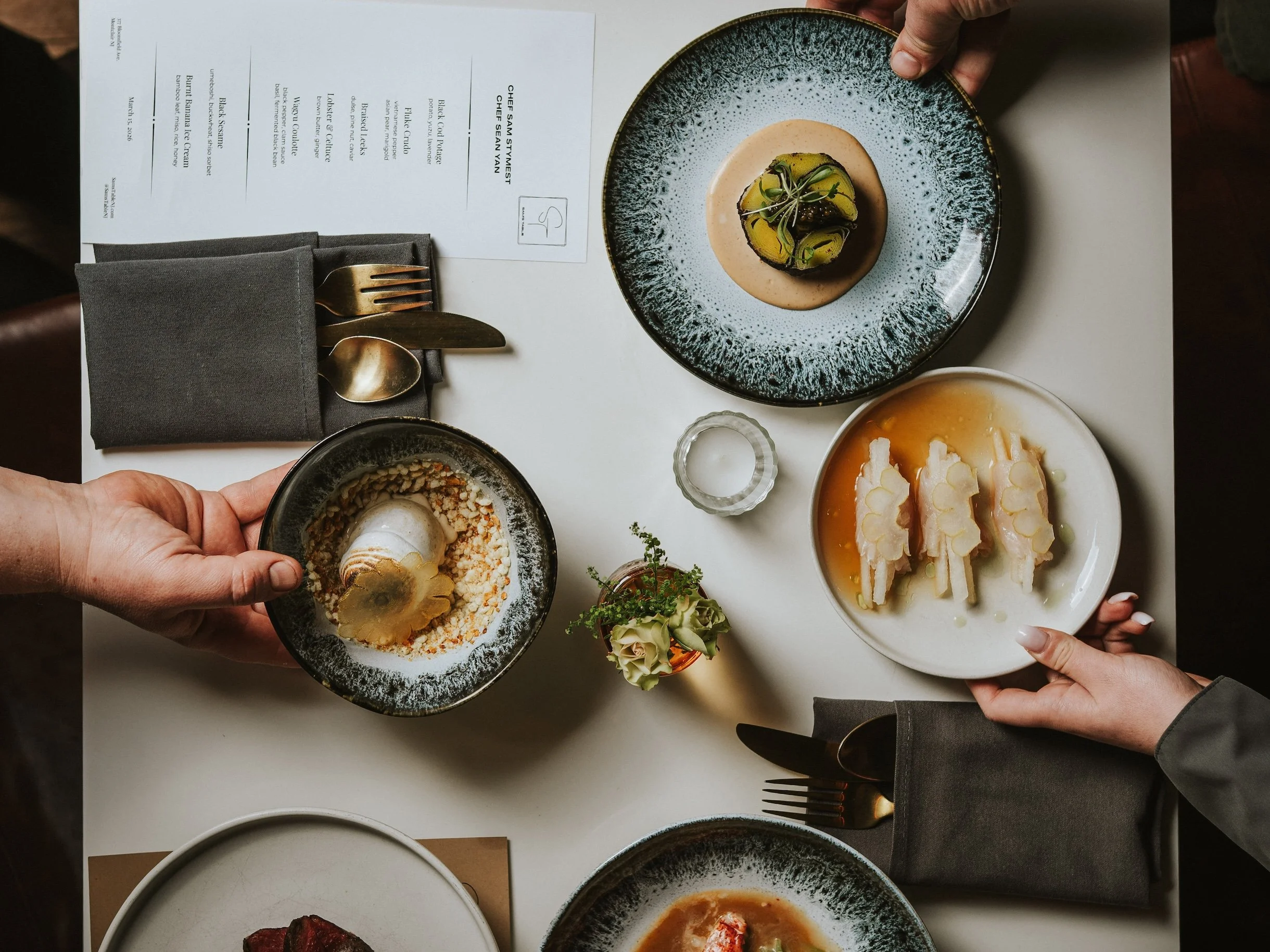 A table with multiple dishes, including a black bowl with a dessert topped with edible gold, a plate with sliced yellow and green vegetables, a bowl of soup, and a bowl of ice cream with a golden garnish, along with a black napkin, fork, and knife.