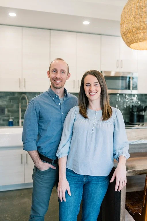 A family of five, including two adults and three children, standing on a roadside with mountains in the background. The man is holding a young girl, and the woman is standing next to two boys. The scene is outdoors in a rural area with grass, trees, and historic wooden barns visible.
