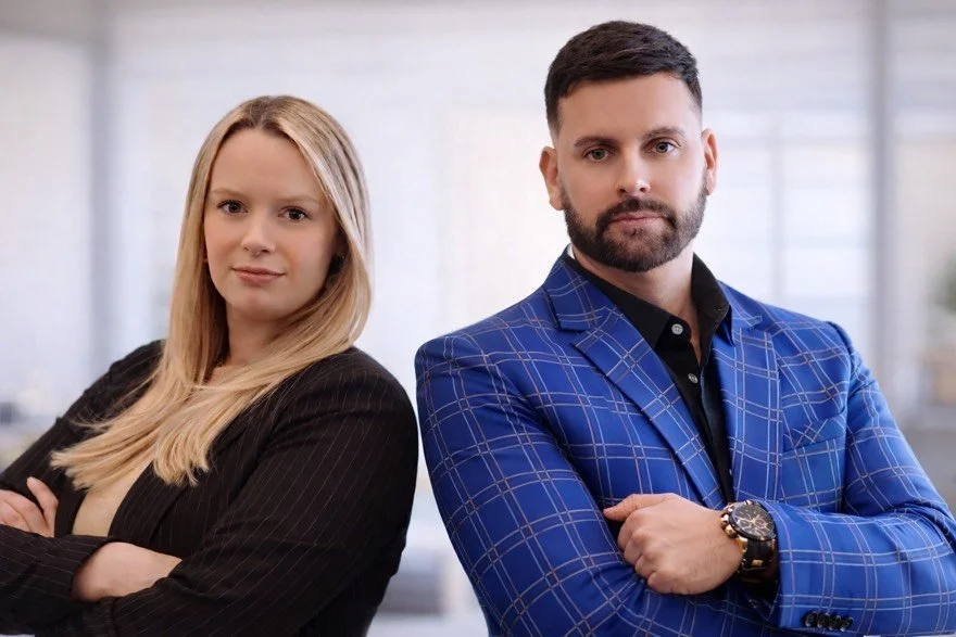 A man and a woman standing with arms crossed, dressed in professional business attire, in a modern office setting.