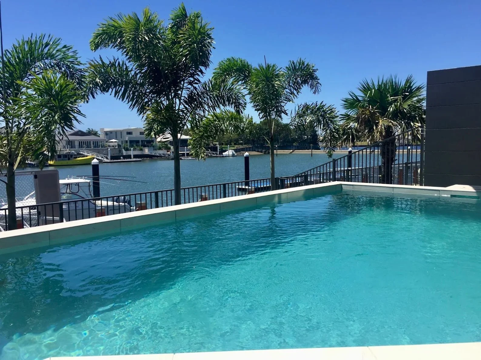 Clear turquoise swimming pool beside a canal, with palm trees and black pool fencing in Bokarina.