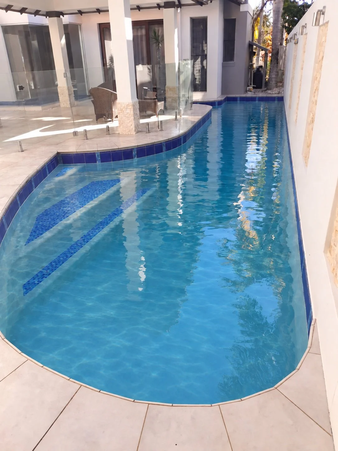 Curved courtyard swimming pool with clear blue water, blue mosaic entry steps and glass fencing beside a covered patio in Bokarina.