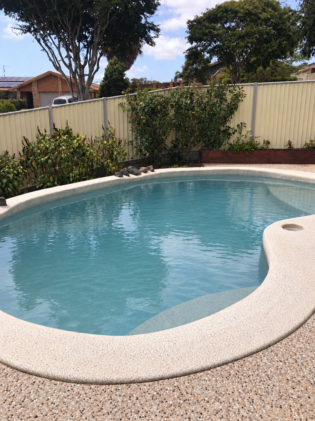 Curved backyard swimming pool in Kunda Park with pale coping, blue water and garden beds along the fence.