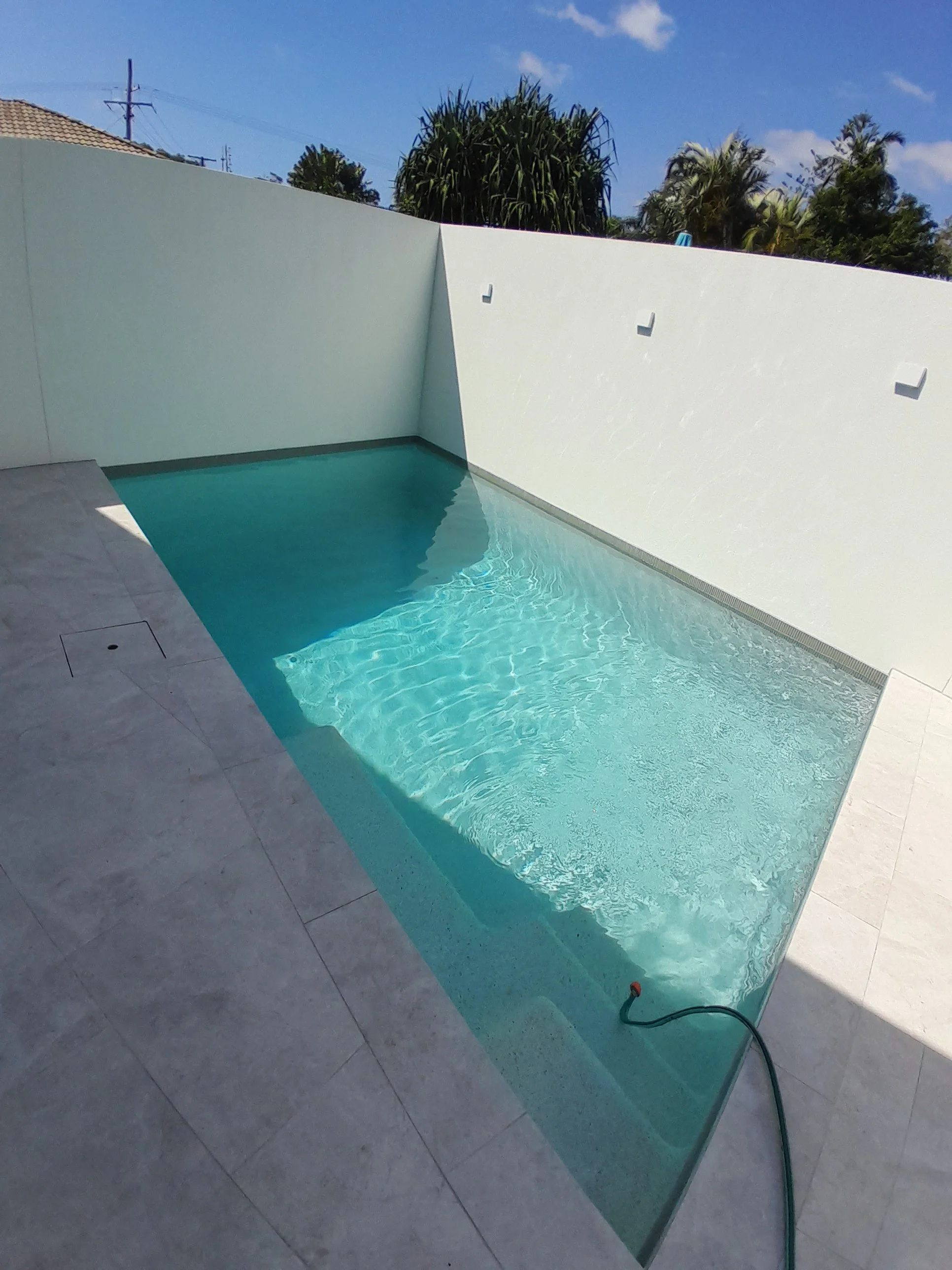 Courtyard pool in Warana with sunlit turquoise water, pale paving, white privacy walls and a pool hose.