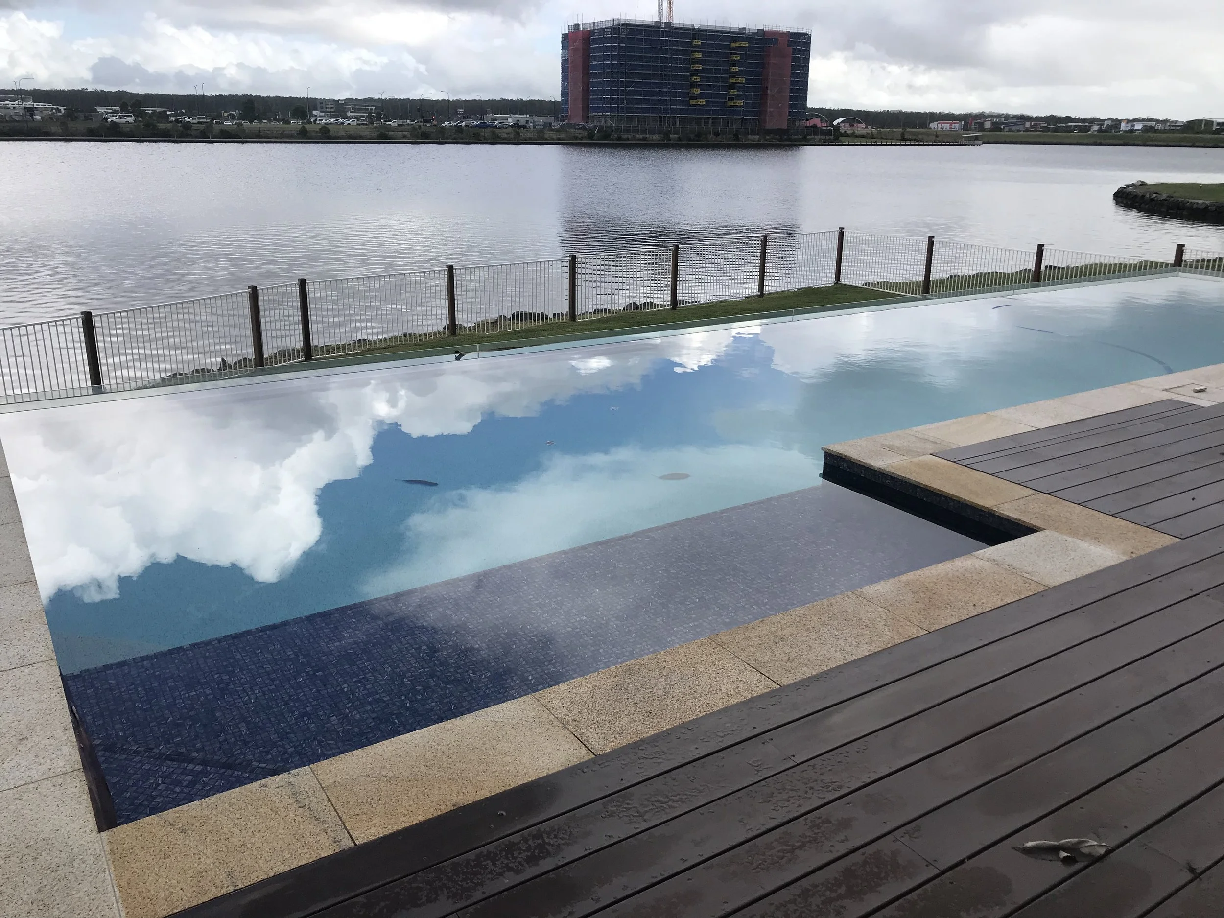 Long lakeside swimming pool reflecting clouds, bordered by timber decking and a glass fence in Bokarina.