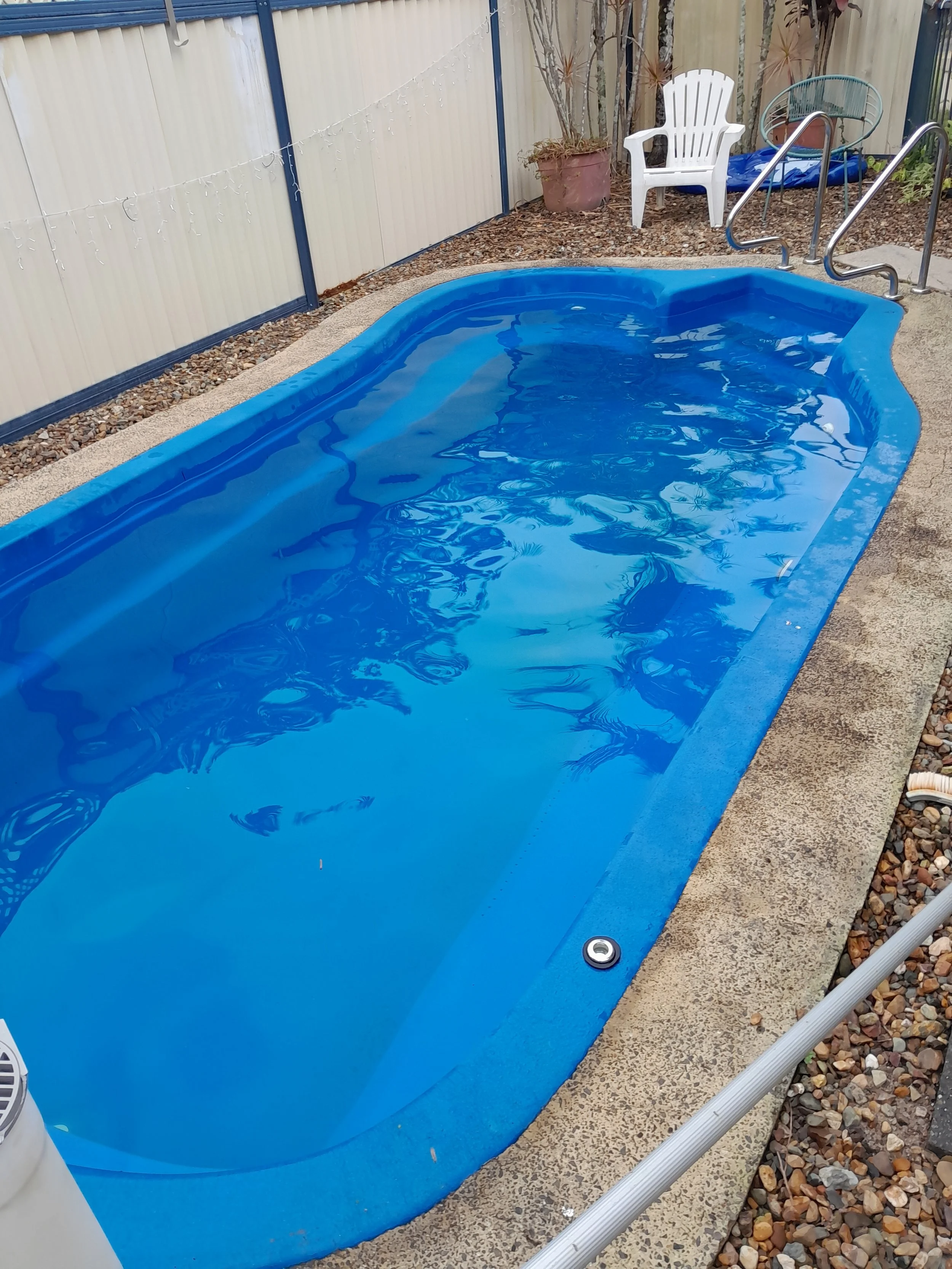 Bright blue backyard swimming pool with curved corners, a handrail and a white chair beside the fence in Parrearra.