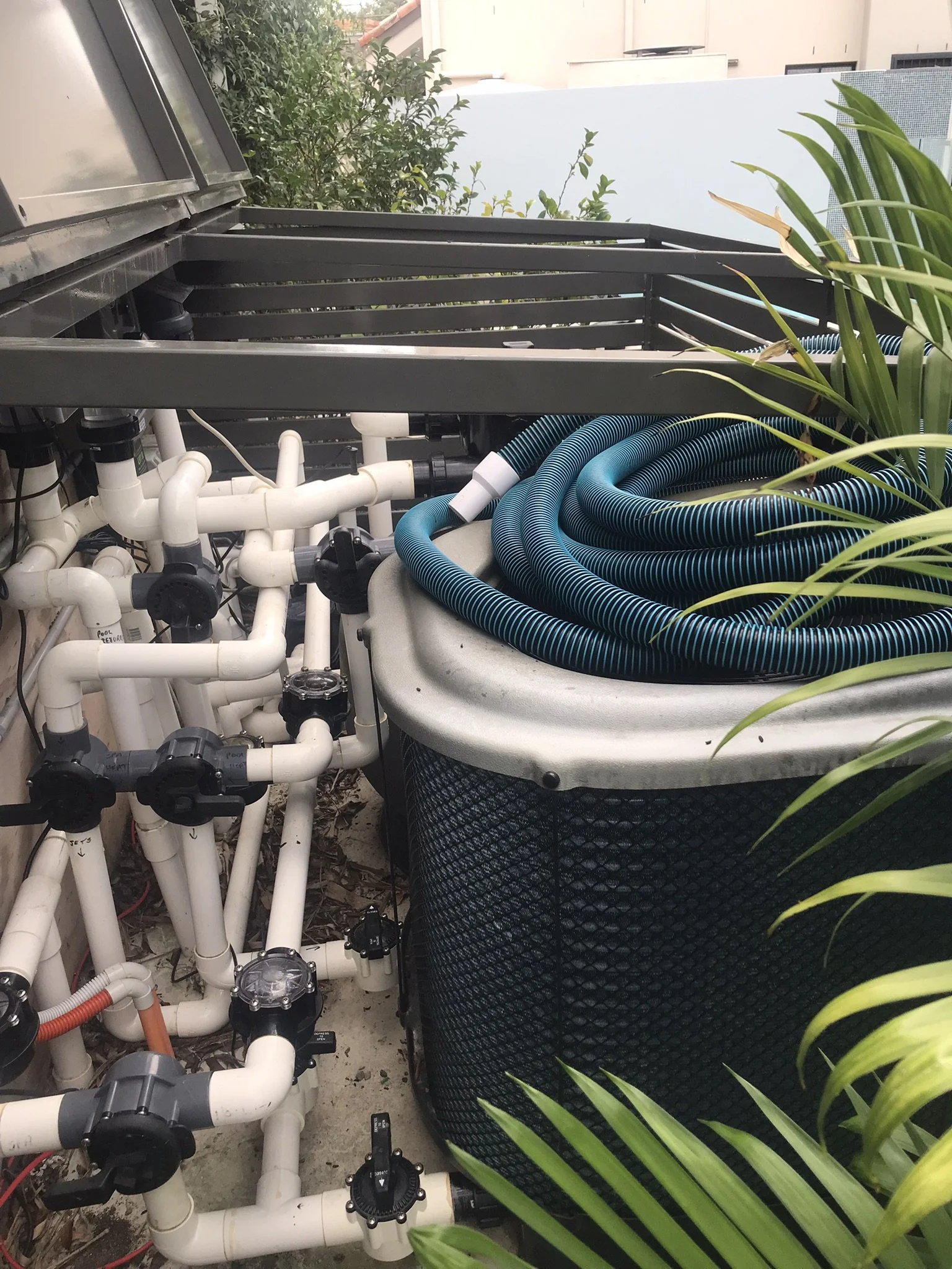 Pool equipment enclosure with white PVC pipes and valves beside a large unit topped with coiled blue hoses.