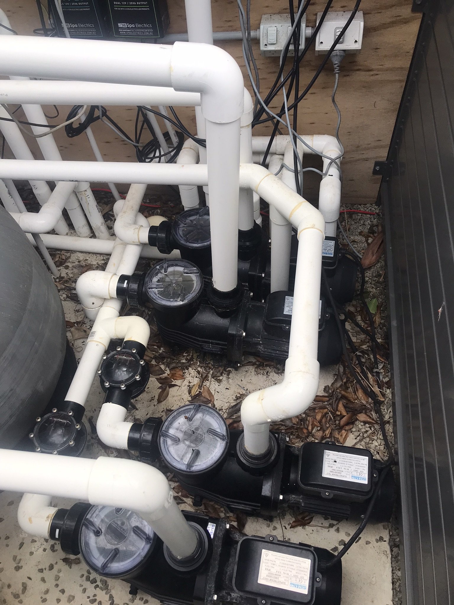 Multiple pool pumps with clear strainer lids connected to closely spaced white PVC pipes inside an equipment enclosure.