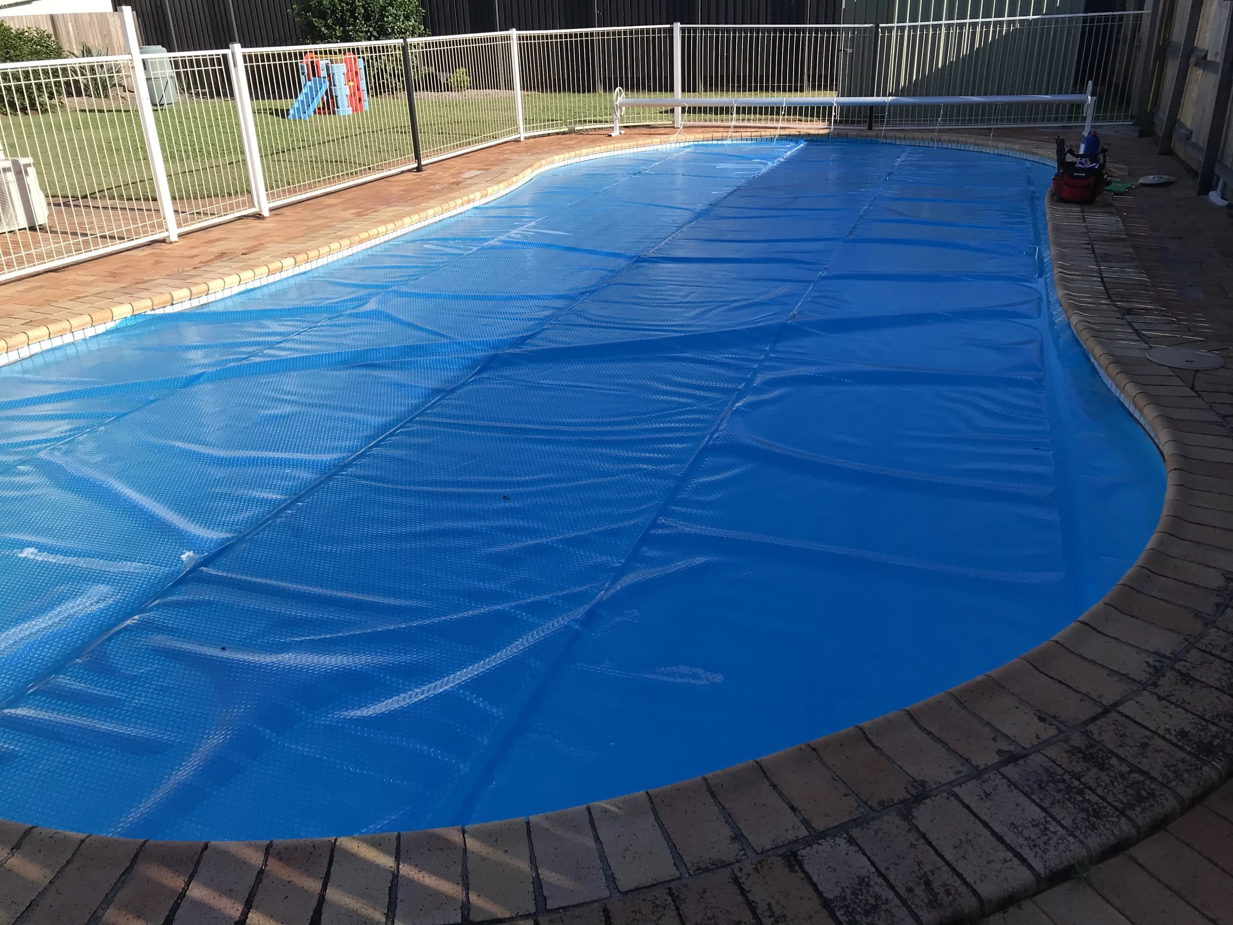 Blue pool cover over a curved swimming pool with brick paving and white pool fencing in Bokarina.