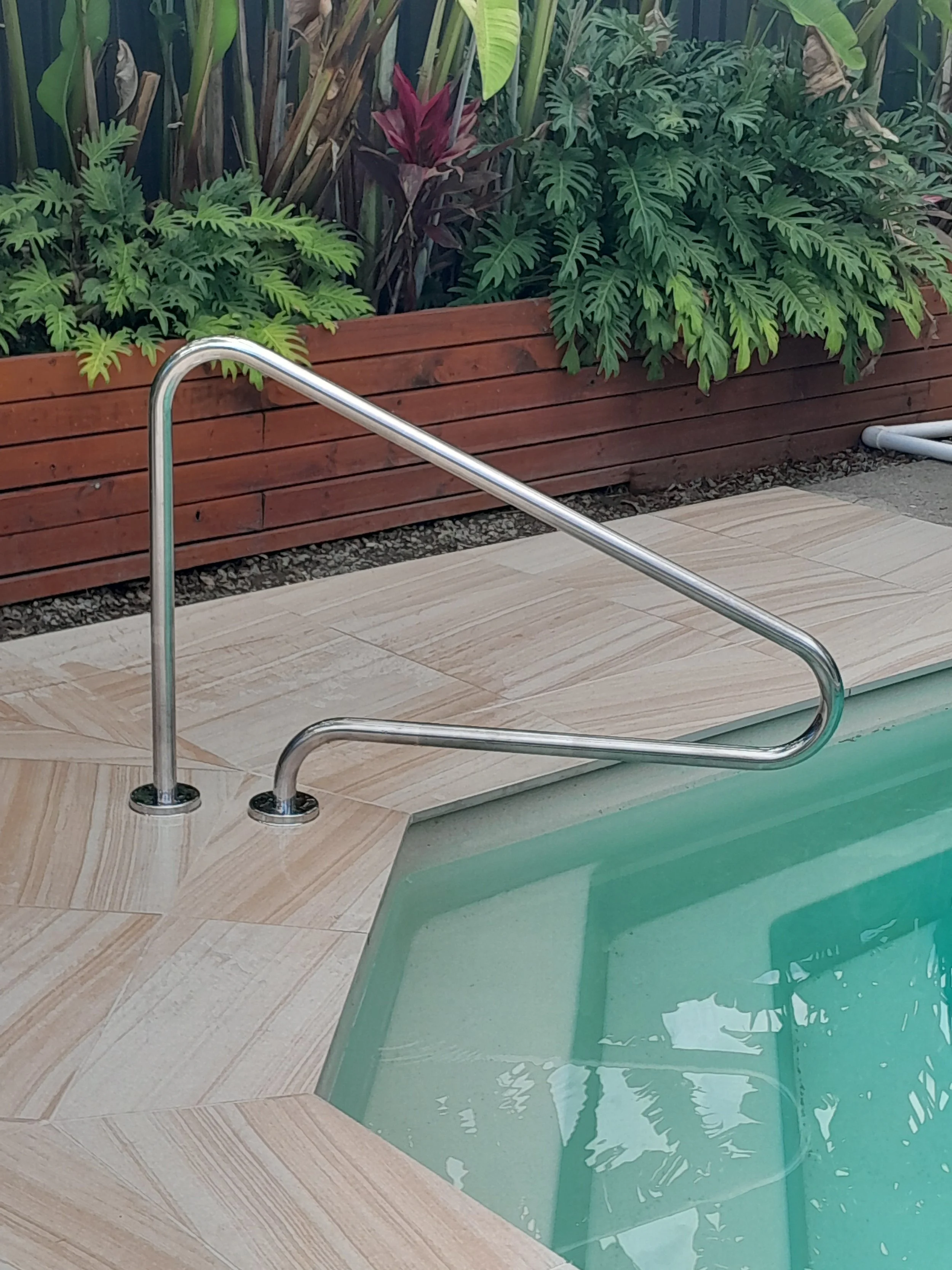Stainless steel handrail beside submerged pool steps, beige paving and a raised garden bed.