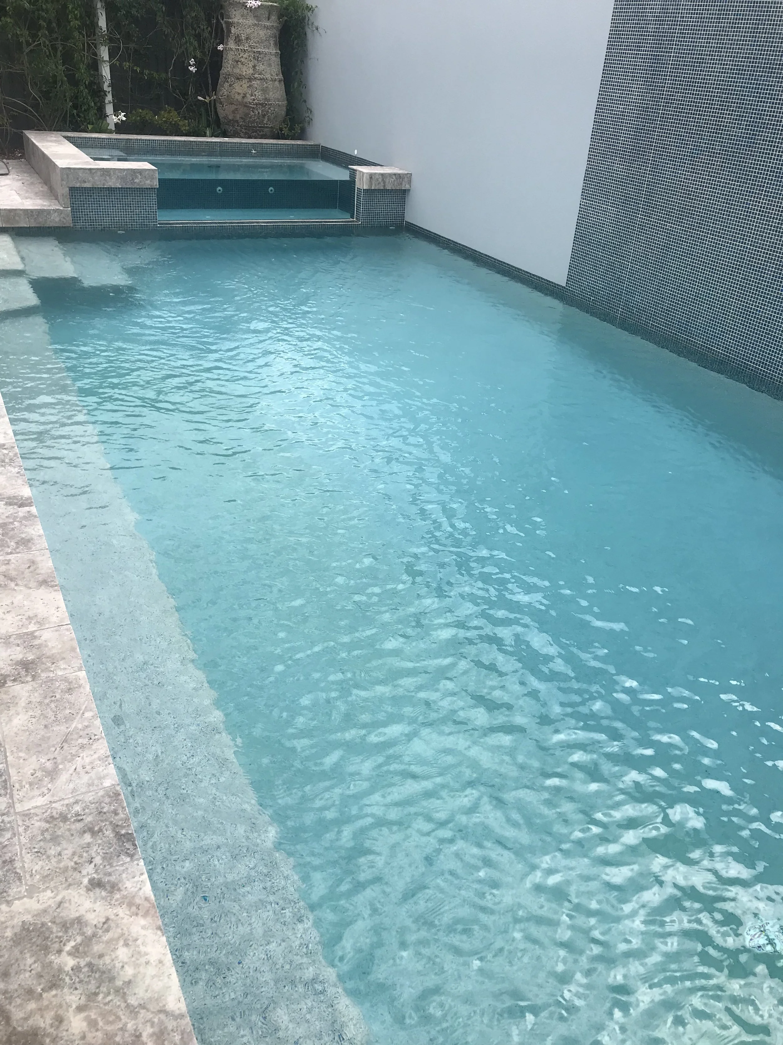 View along a rectangular swimming pool towards a raised spa, bordered by stone coping and a high wall.