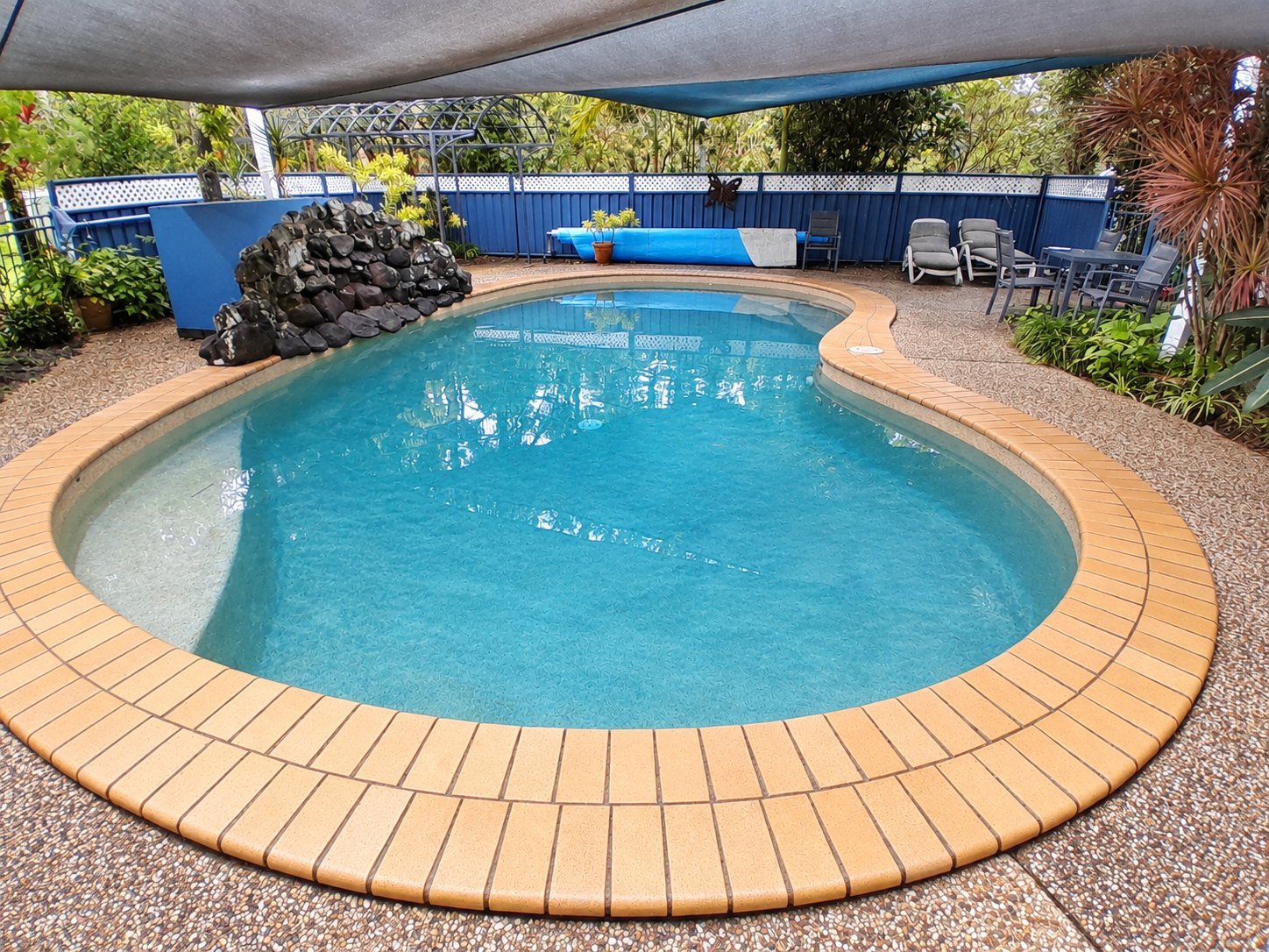 Curved swimming pool beneath shade sails with a rock feature in Ilkley.